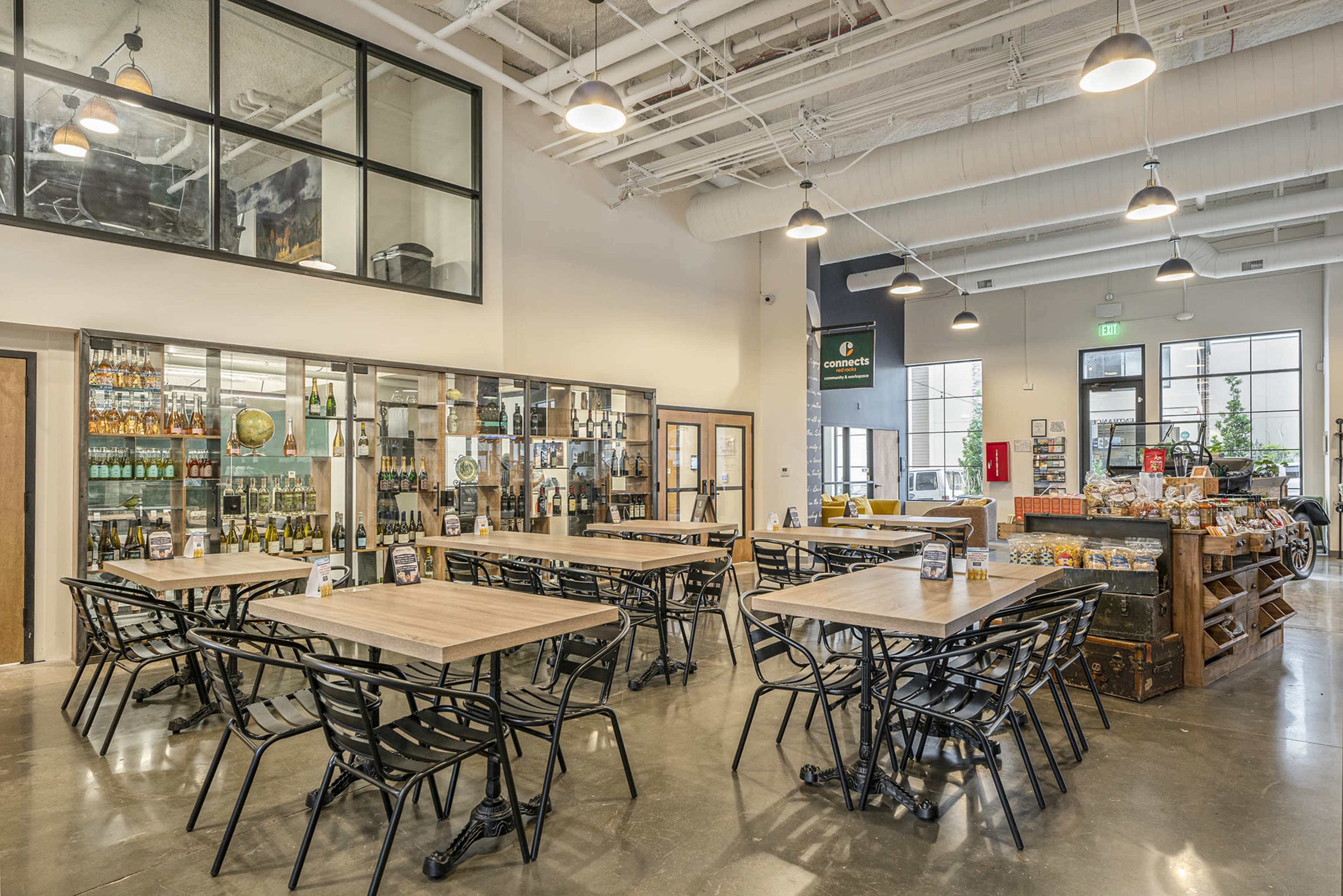 The image shows a spacious, modern dining area with multiple wooden tables and black chairs, as well as a display case filled with various bottled products.
