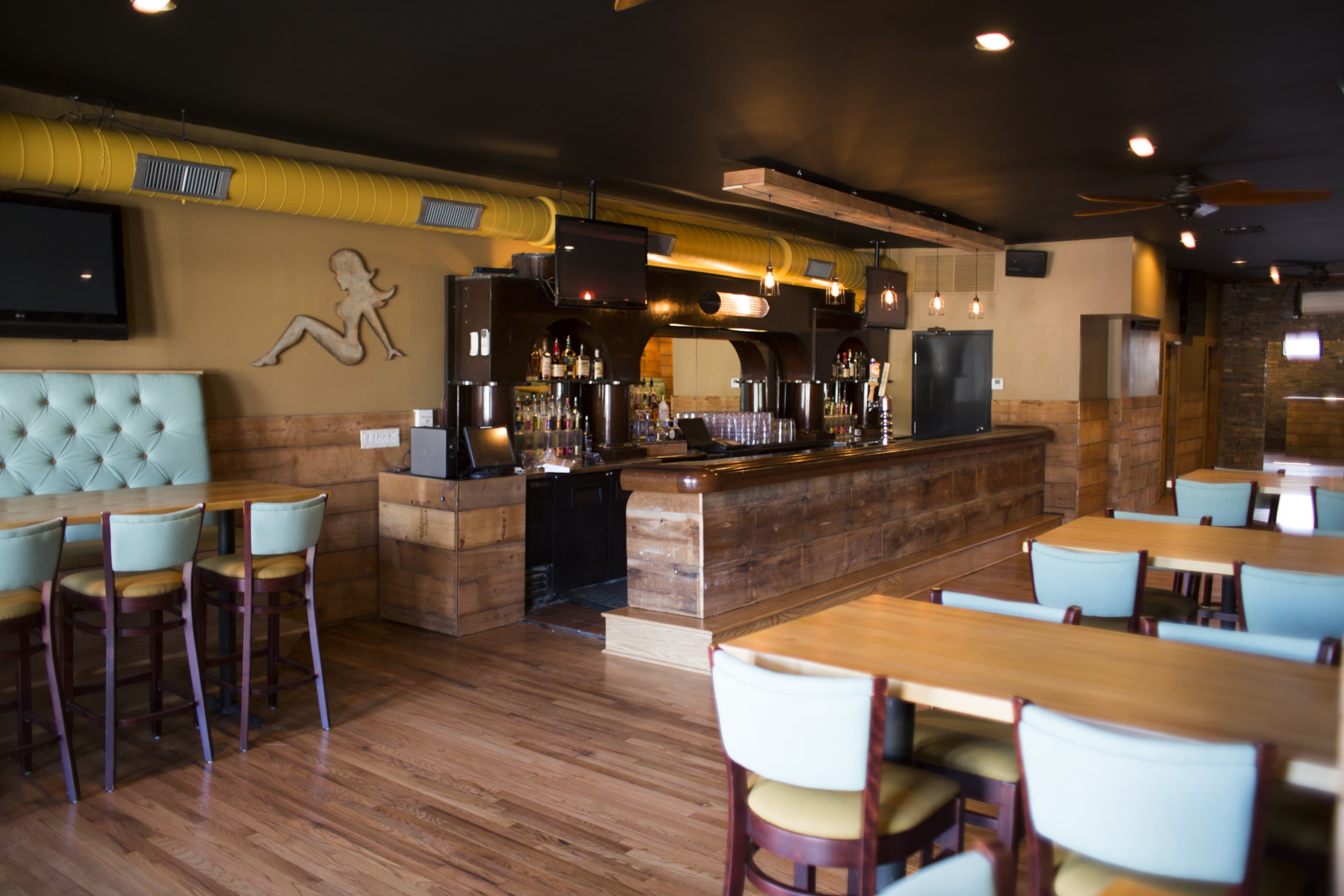 The image shows an interior of a bar and restaurant with wooden furnishings, a long bar counter, and table seating arranged throughout the space.