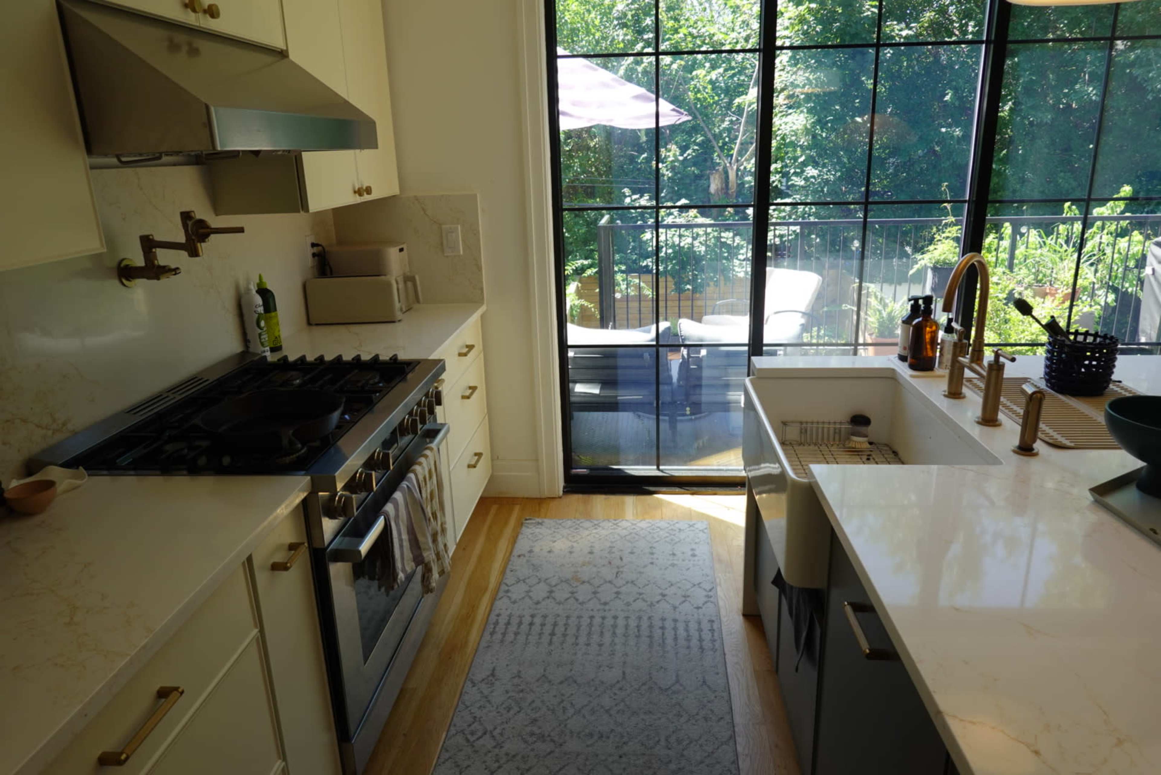 A modern kitchen features a stainless steel oven and stove, white countertops, and a large window overlooking a garden area with outdoor seating.