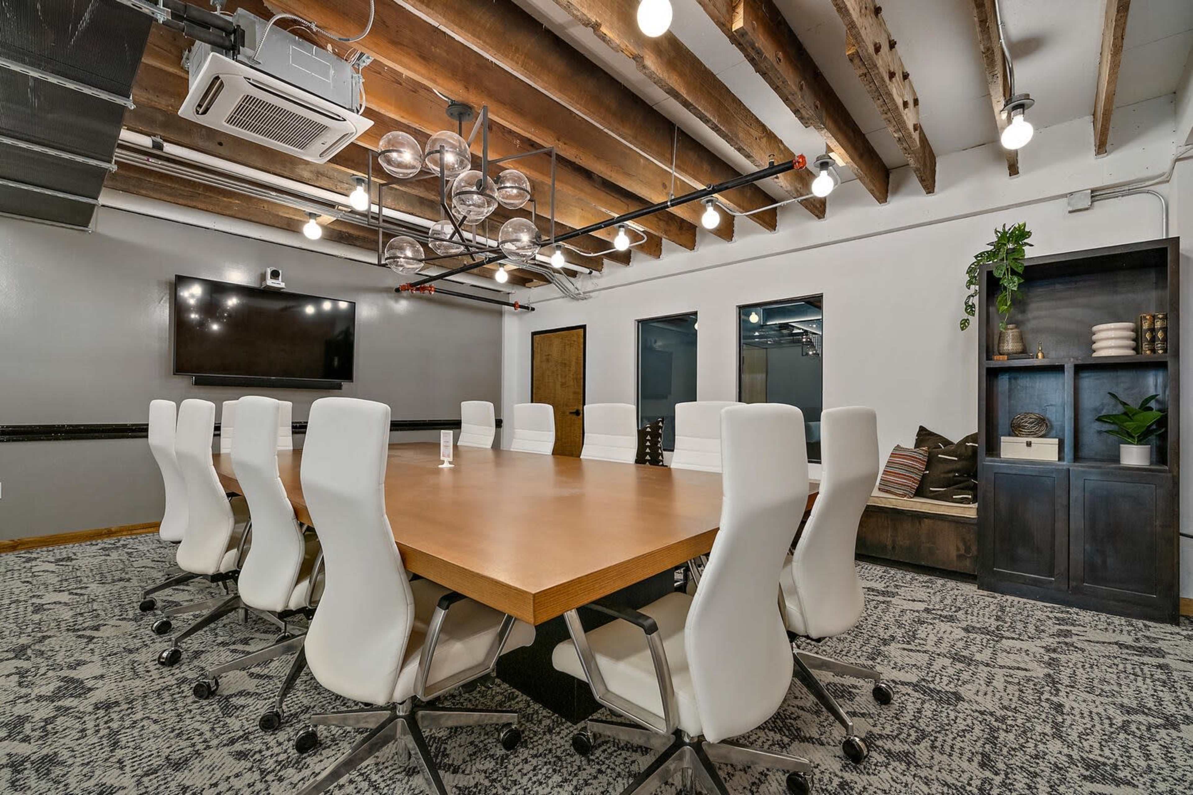 A conference room features a large wooden table surrounded by white chairs, with modern lighting and a television on the wall.