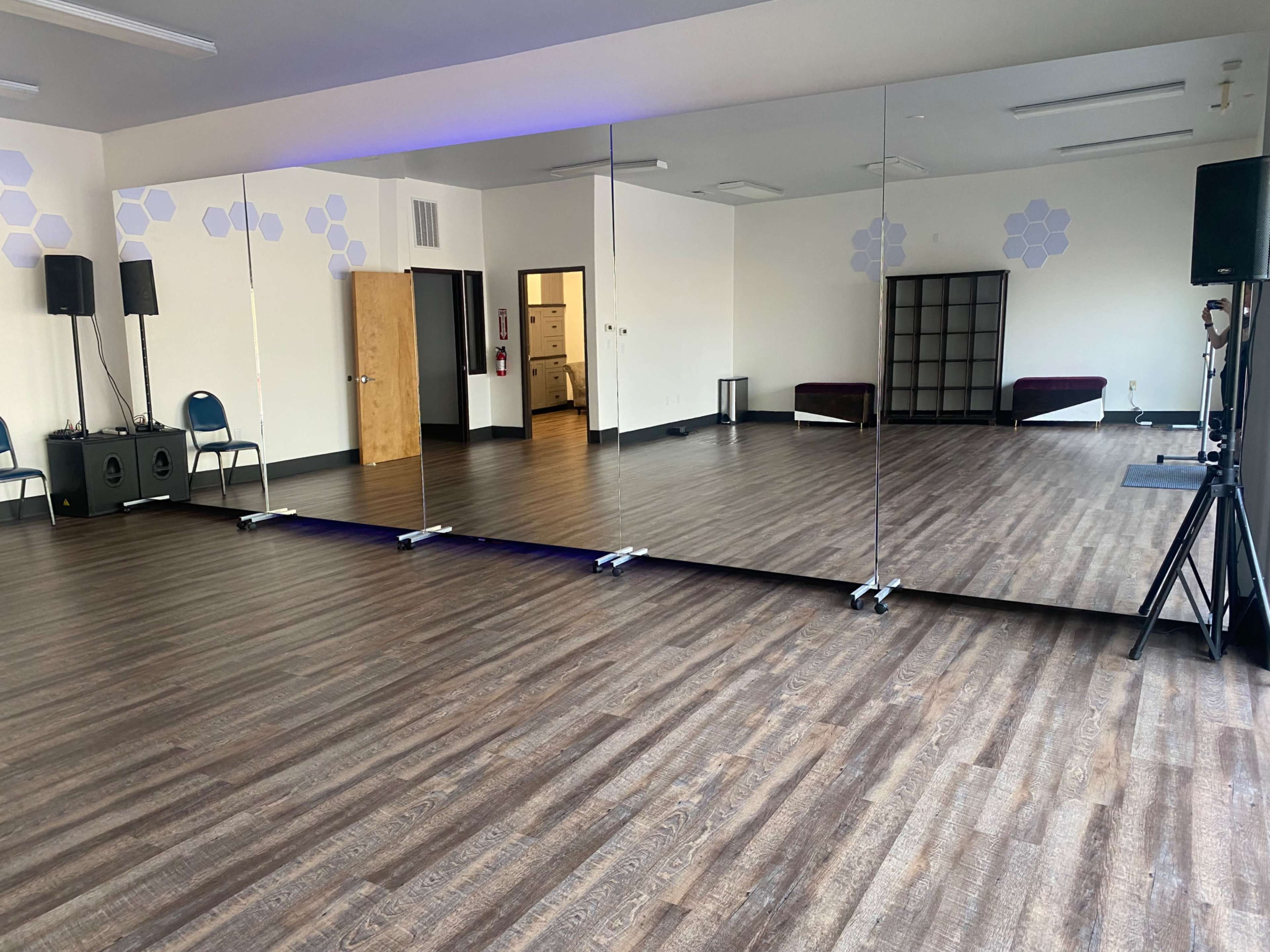 The image showcases a spacious dance studio featuring a mirrored wall, wooden flooring, and simple chairs along the sides.