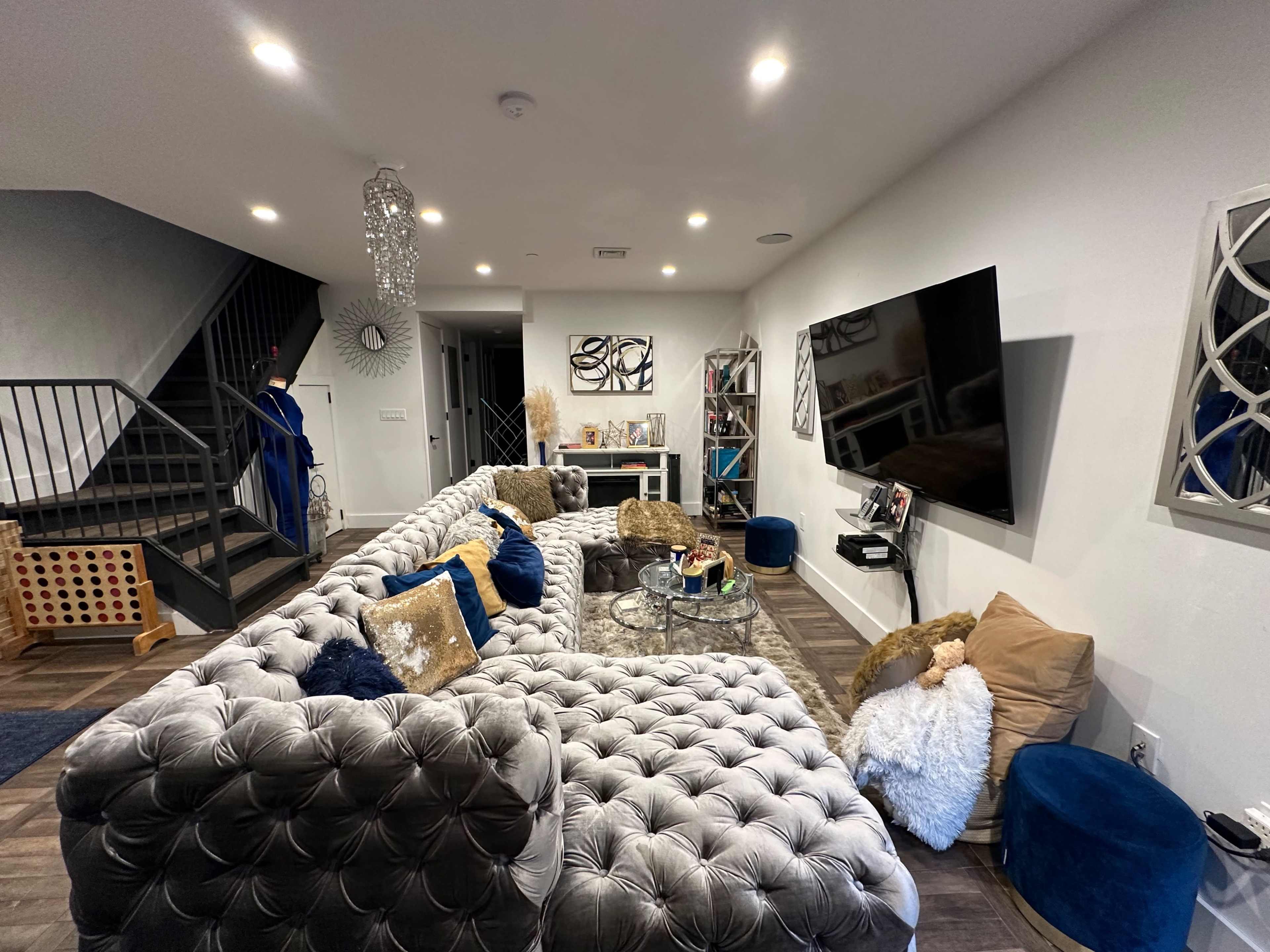A modern living room features a large tufted sectional sofa, a coffee table, a wall-mounted television, and decorative elements along with a staircase in the background.