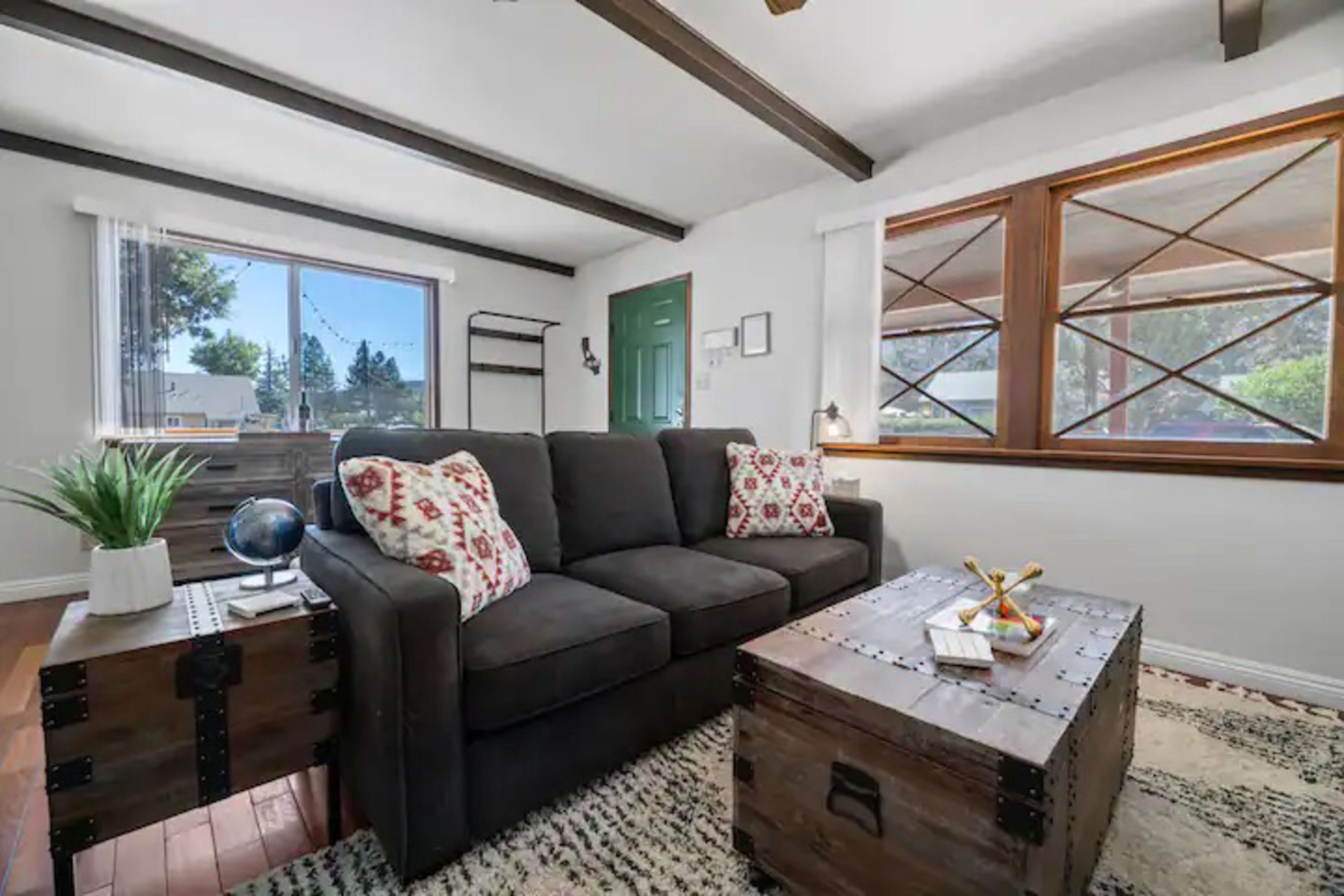 A living room featuring a dark couch with patterned pillows, a wooden coffee table, and large windows that allow natural light to enter the space.