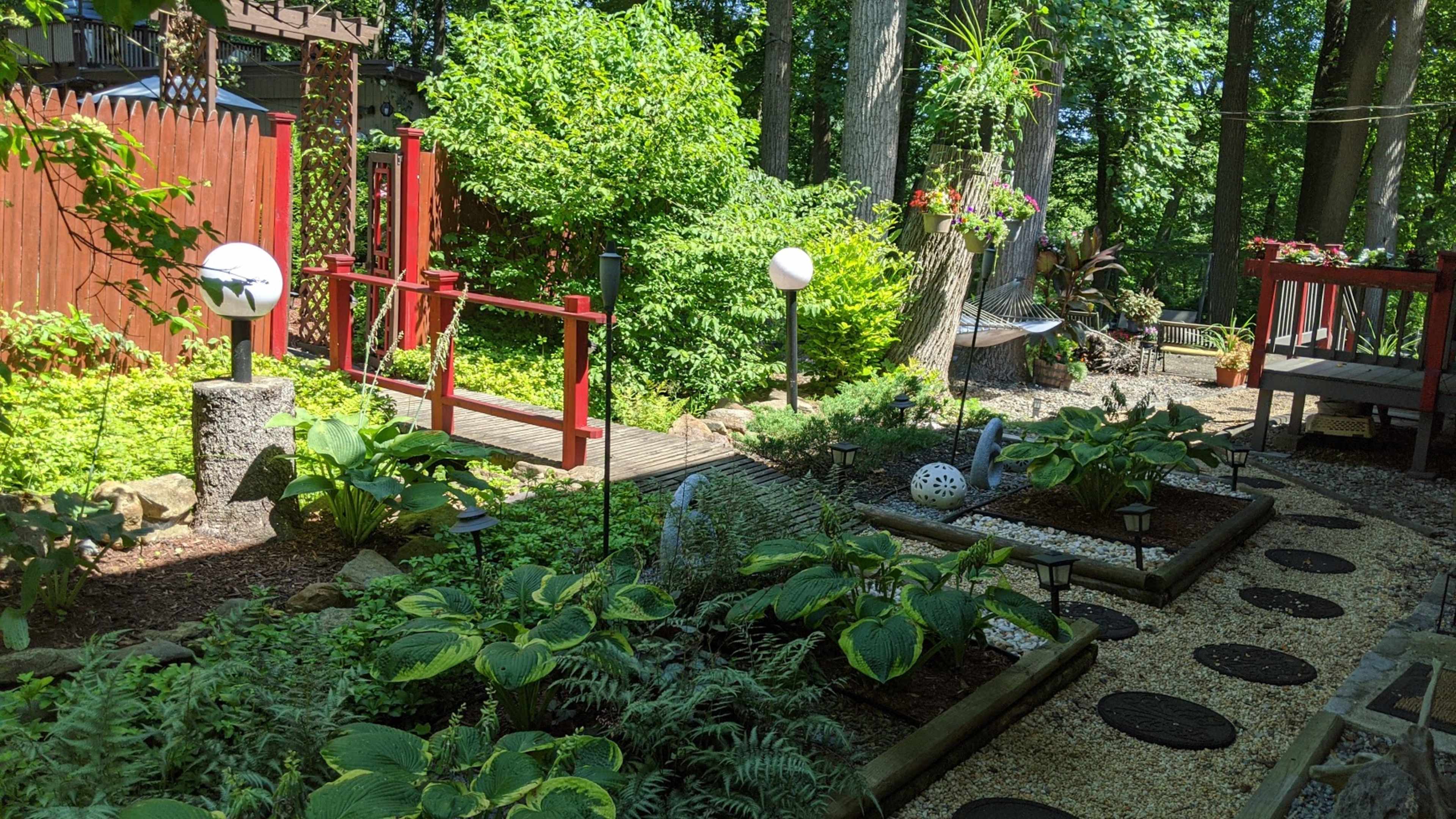 A landscaped garden with a wooden bridge, various plants, and decorative lighting elements amidst trees.