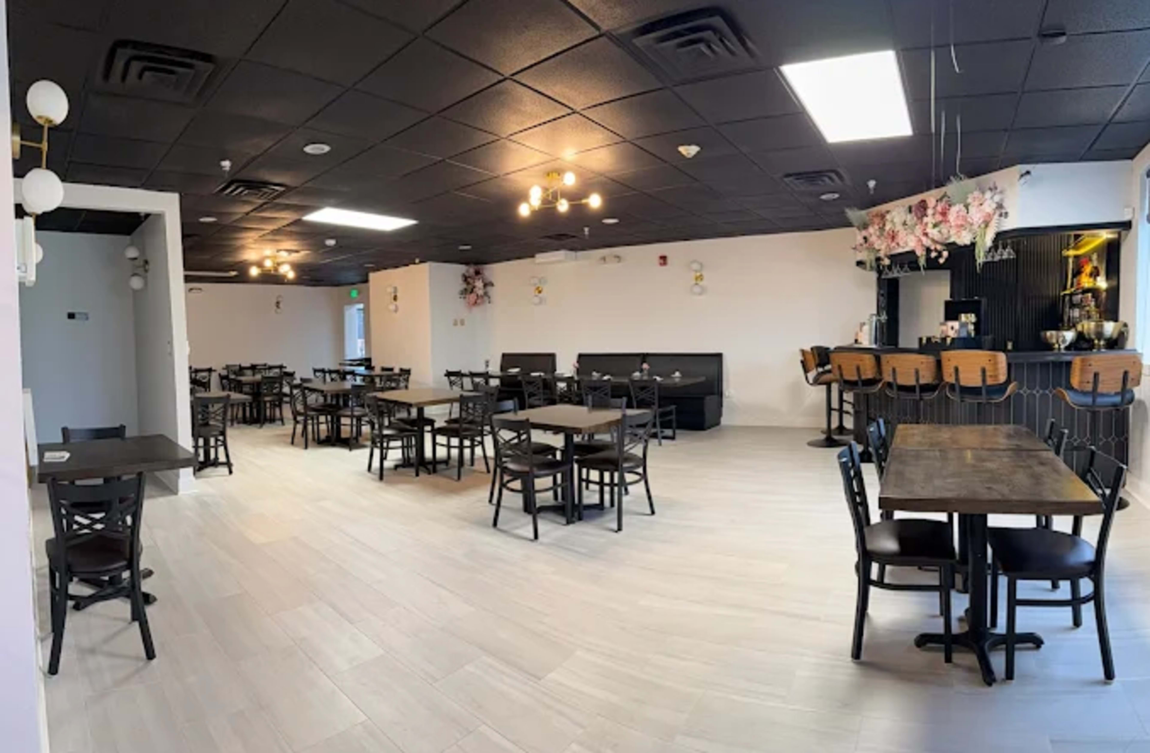 The image shows a spacious restaurant interior with black tables and chairs arranged throughout, featuring a bar area in the back surrounded by decorative elements.