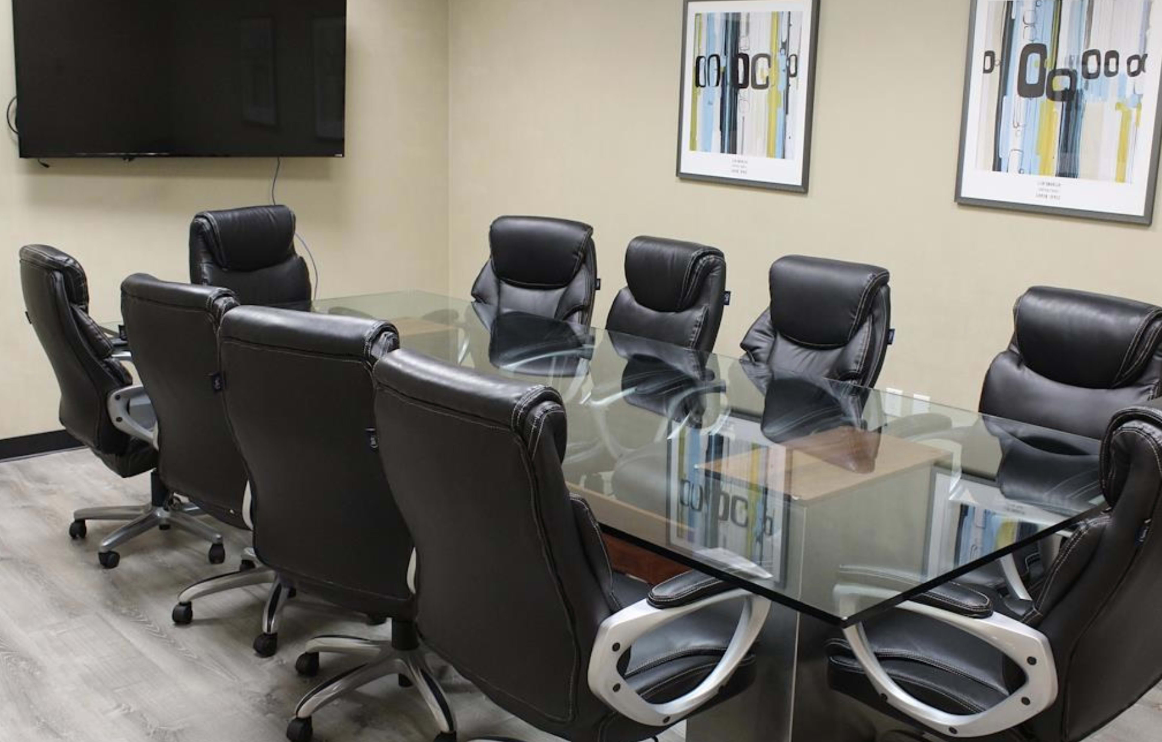 A modern conference room features a long glass table surrounded by eight black leather chairs, with a television screen and framed artwork on the walls.