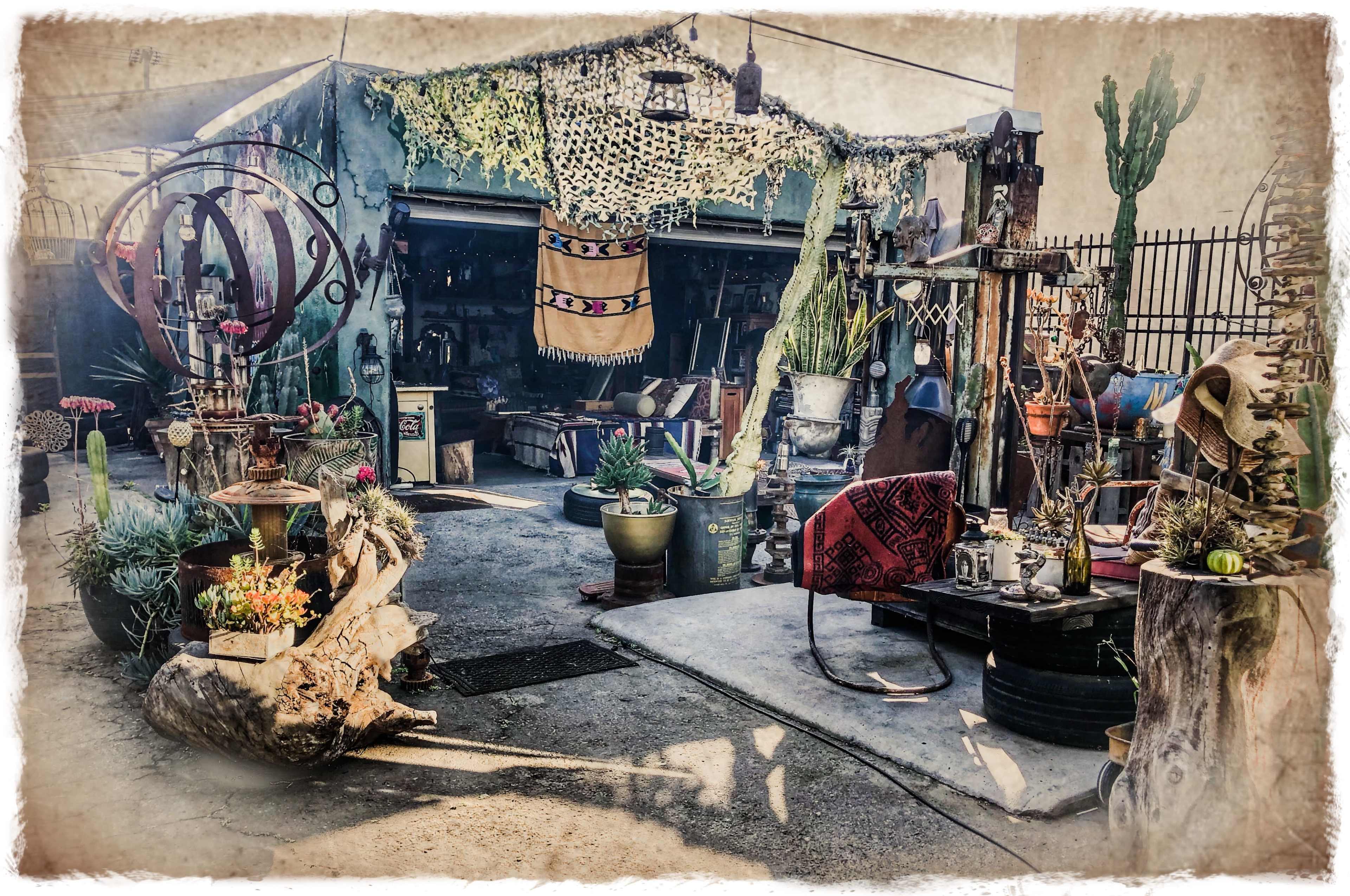 A cluttered outdoor market space filled with various plants, decorative items, and rustic furniture under a canopy adorned with textiles.