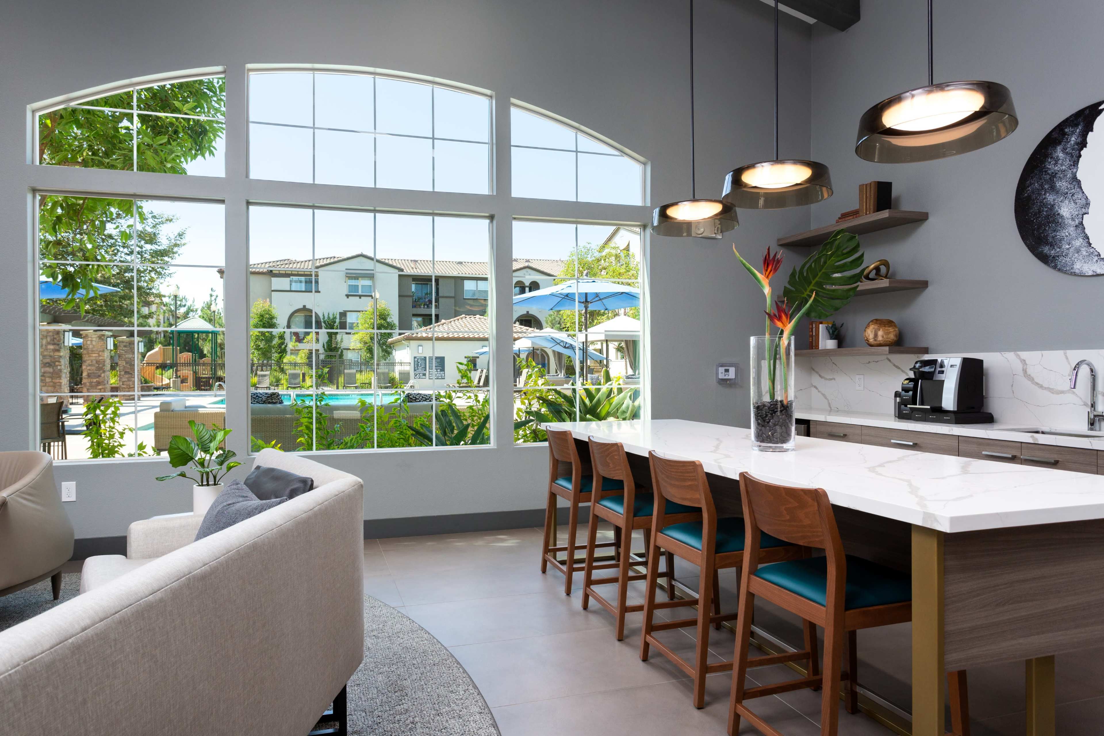 A modern kitchen area with a marble countertop and wooden bar stools, featuring large windows that provide a view of a pool and outdoor space.