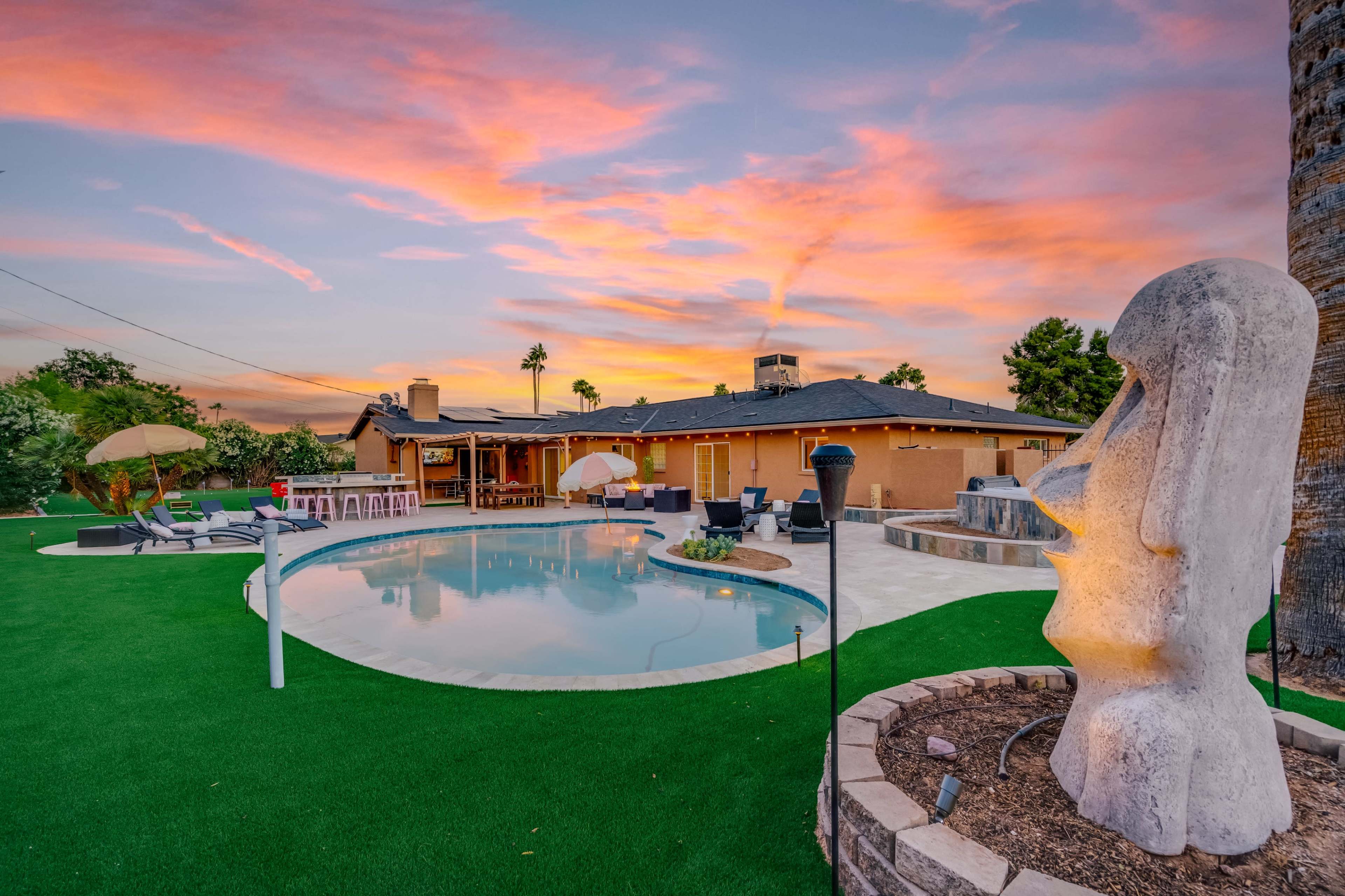 A modern backyard with a swimming pool, landscaping, lounge chairs, and a large decorative sculpture, set against a colorful sunset sky.