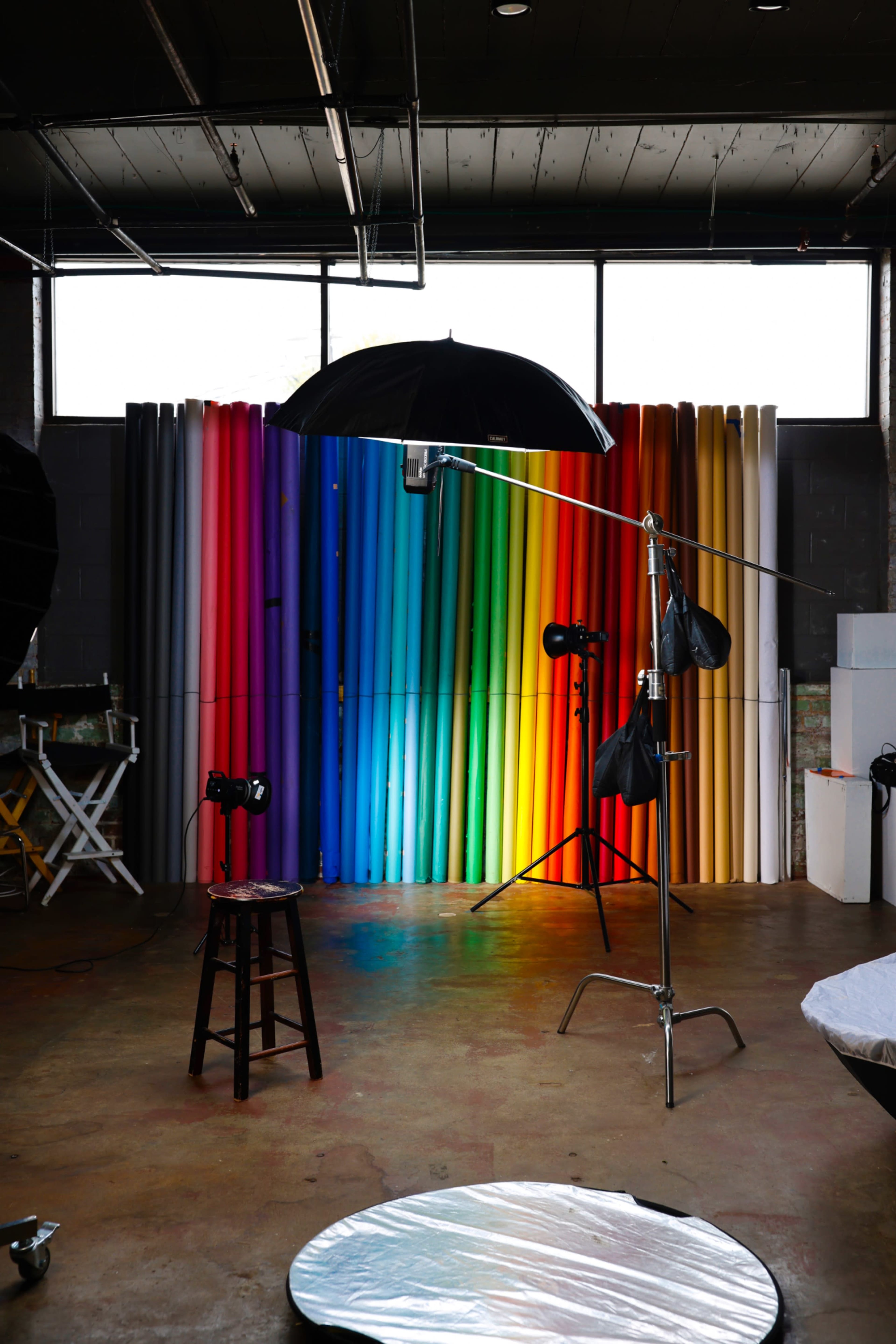 A photography studio features assorted colorful backdrops, equipment like lights and umbrellas, and a stool in the foreground.