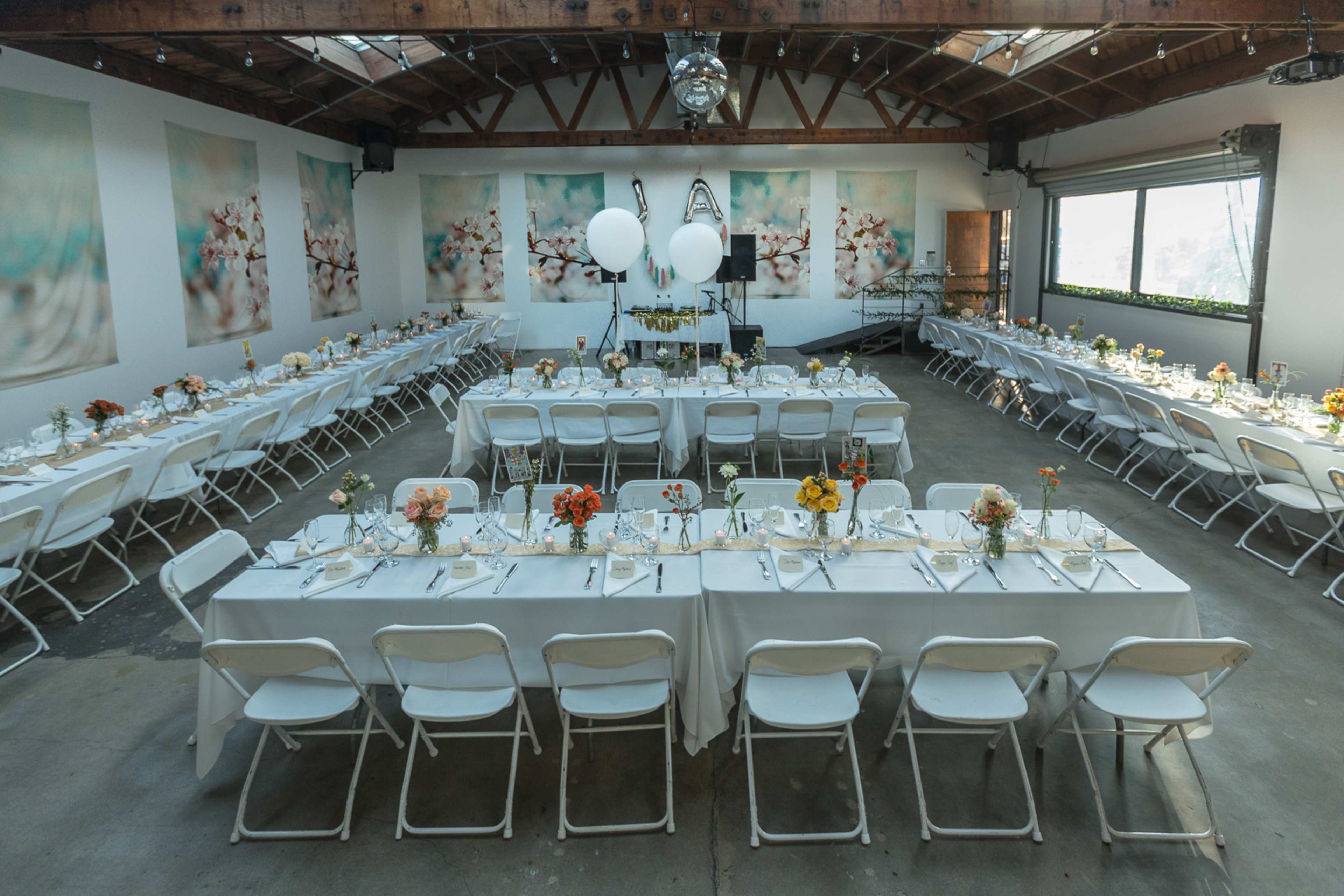 A large event space is arranged with two long tables set for a meal, adorned with floral centerpieces and white chairs, against a backdrop of floral artwork.