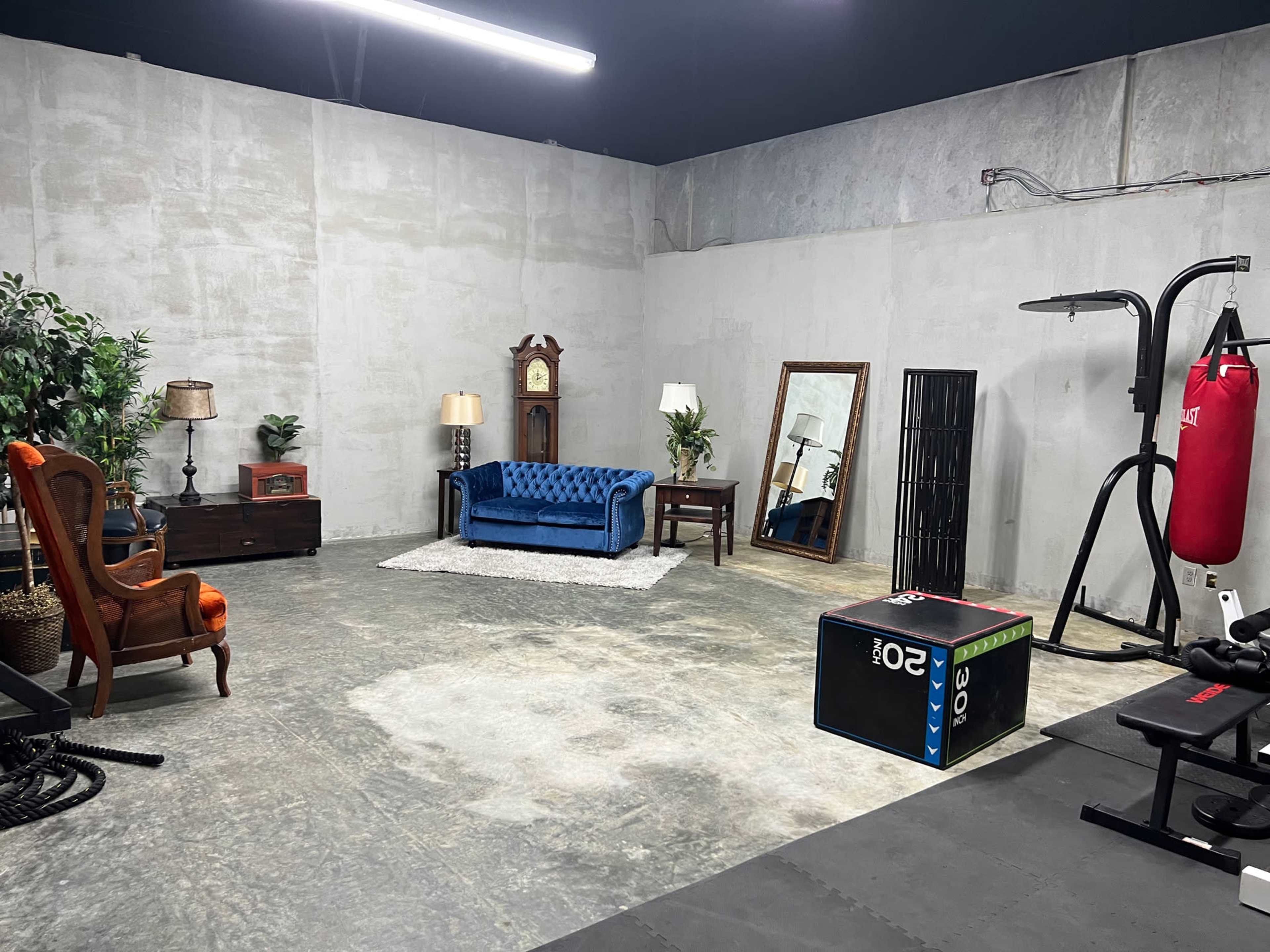 The image shows an empty gym interior with various pieces of furniture, including a blue sofa, an ornate clock, and exercise equipment like a punching bag and weights.