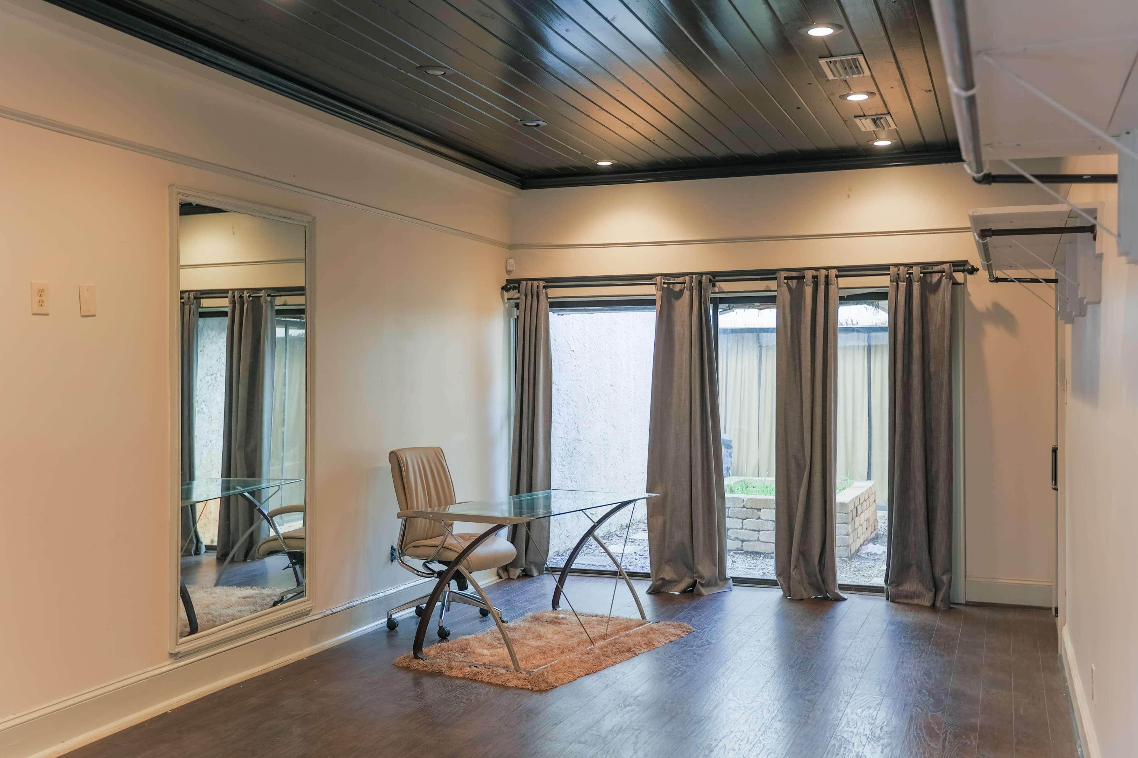 A room with a glass desk and chair, a mirror on one wall, and sliding curtains covering a glass door leading to an outdoor area.