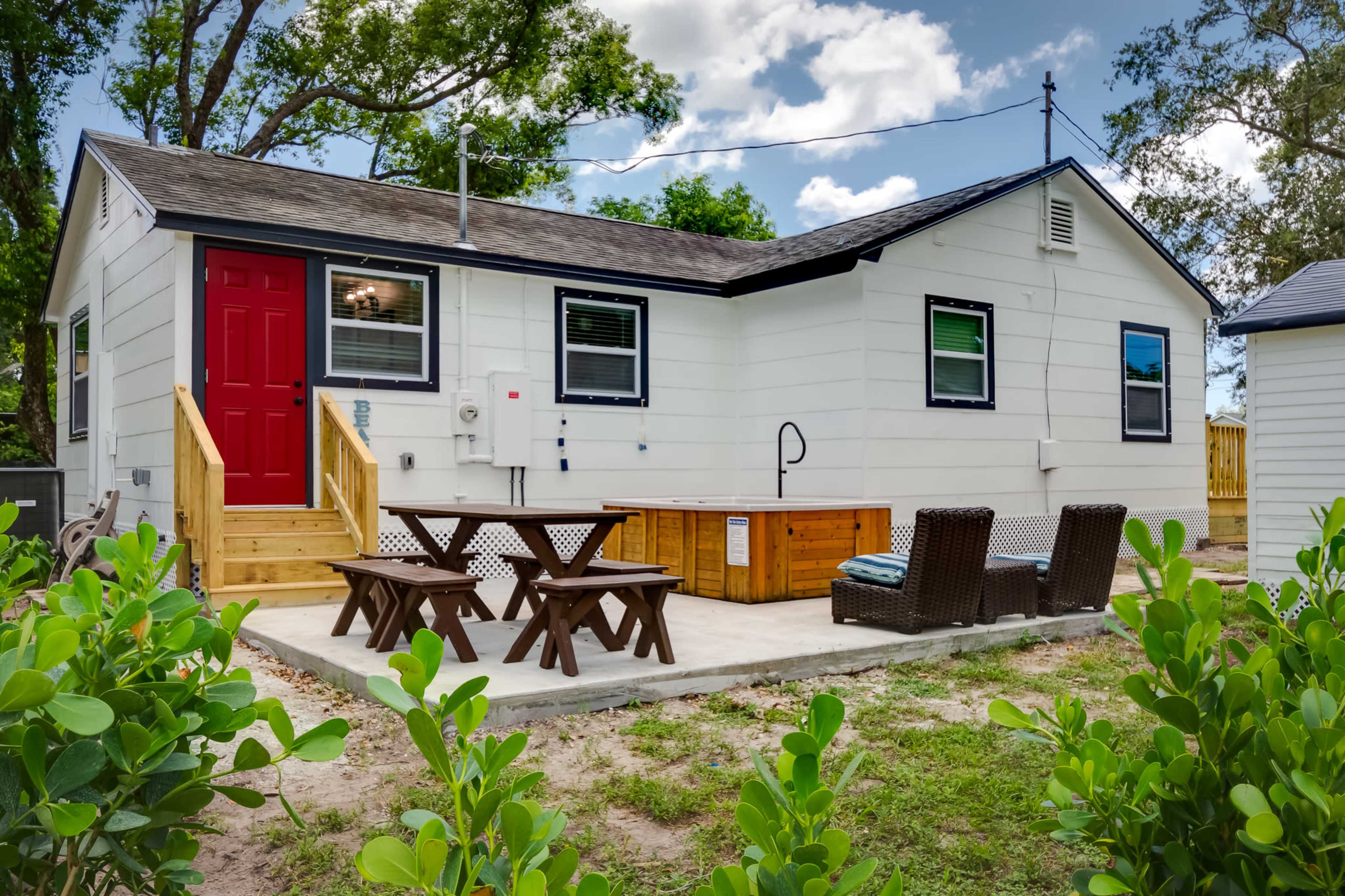 A small white house with a red door features a wooden deck, a hot tub, and outdoor seating surrounded by greenery.