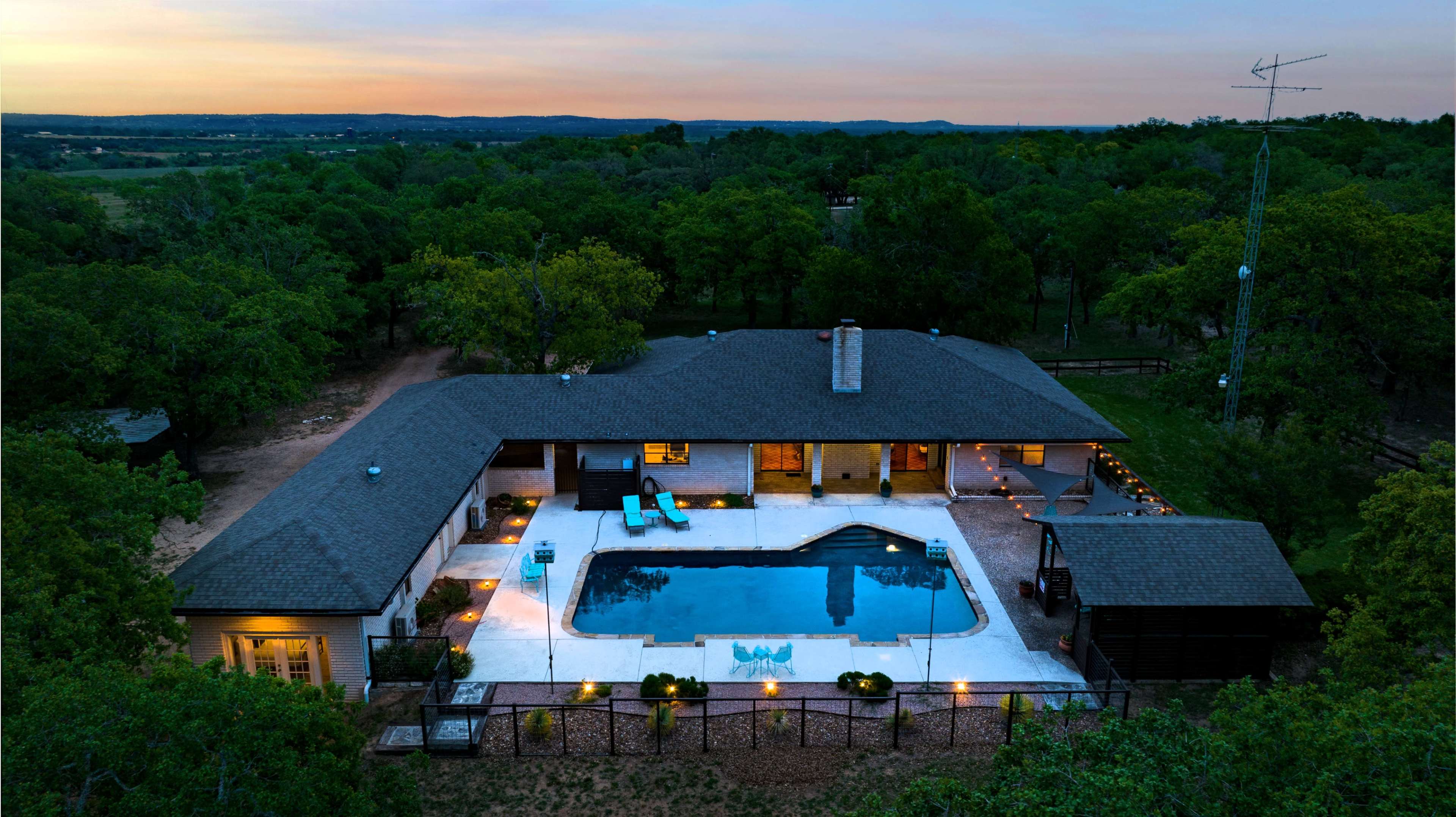 A single-story house with a swimming pool surrounded by a patio and landscaped yard, set against a background of trees during sunset.