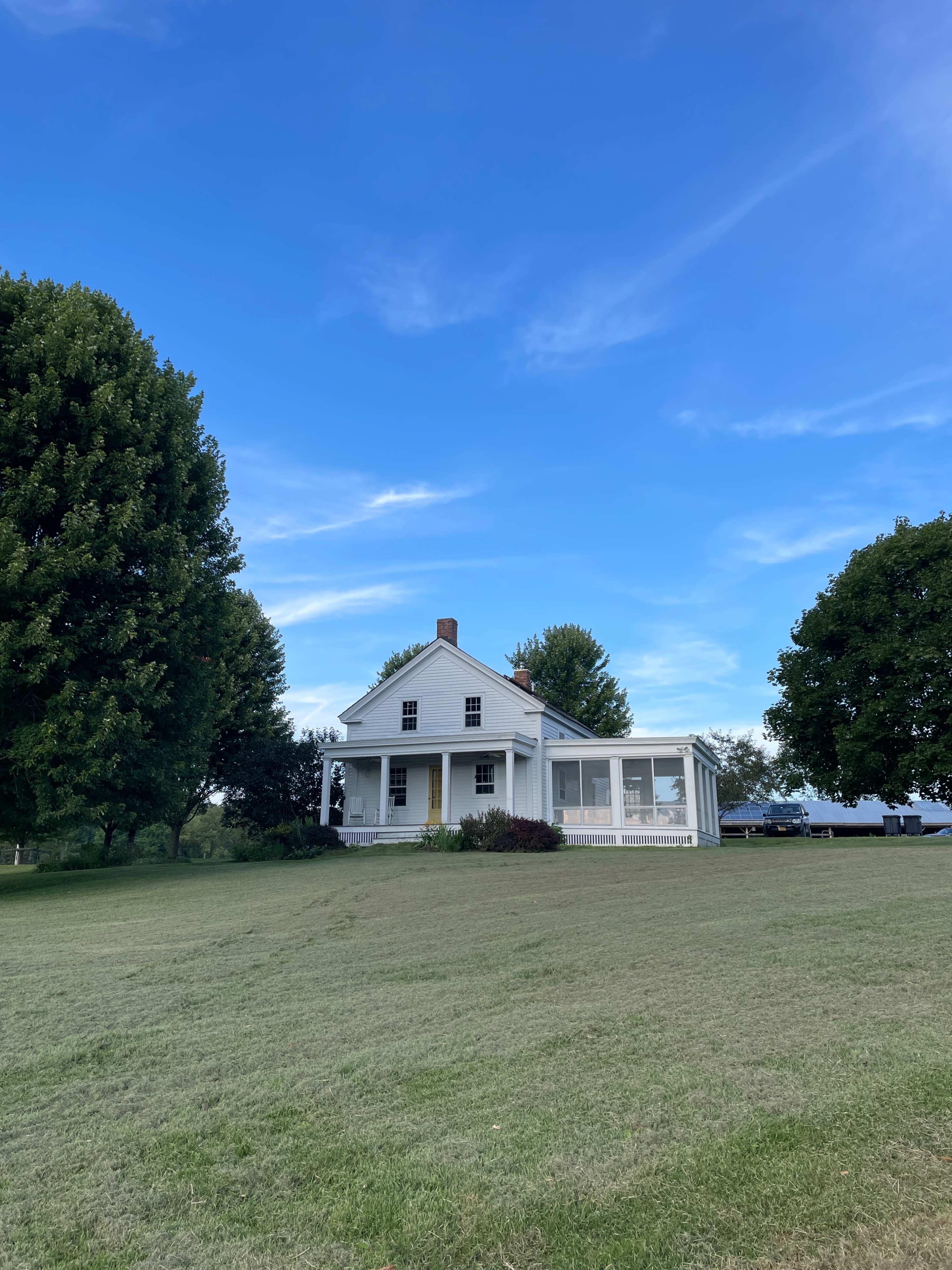 A white two-story house with a porch is surrounded by green grass and trees under a blue sky.