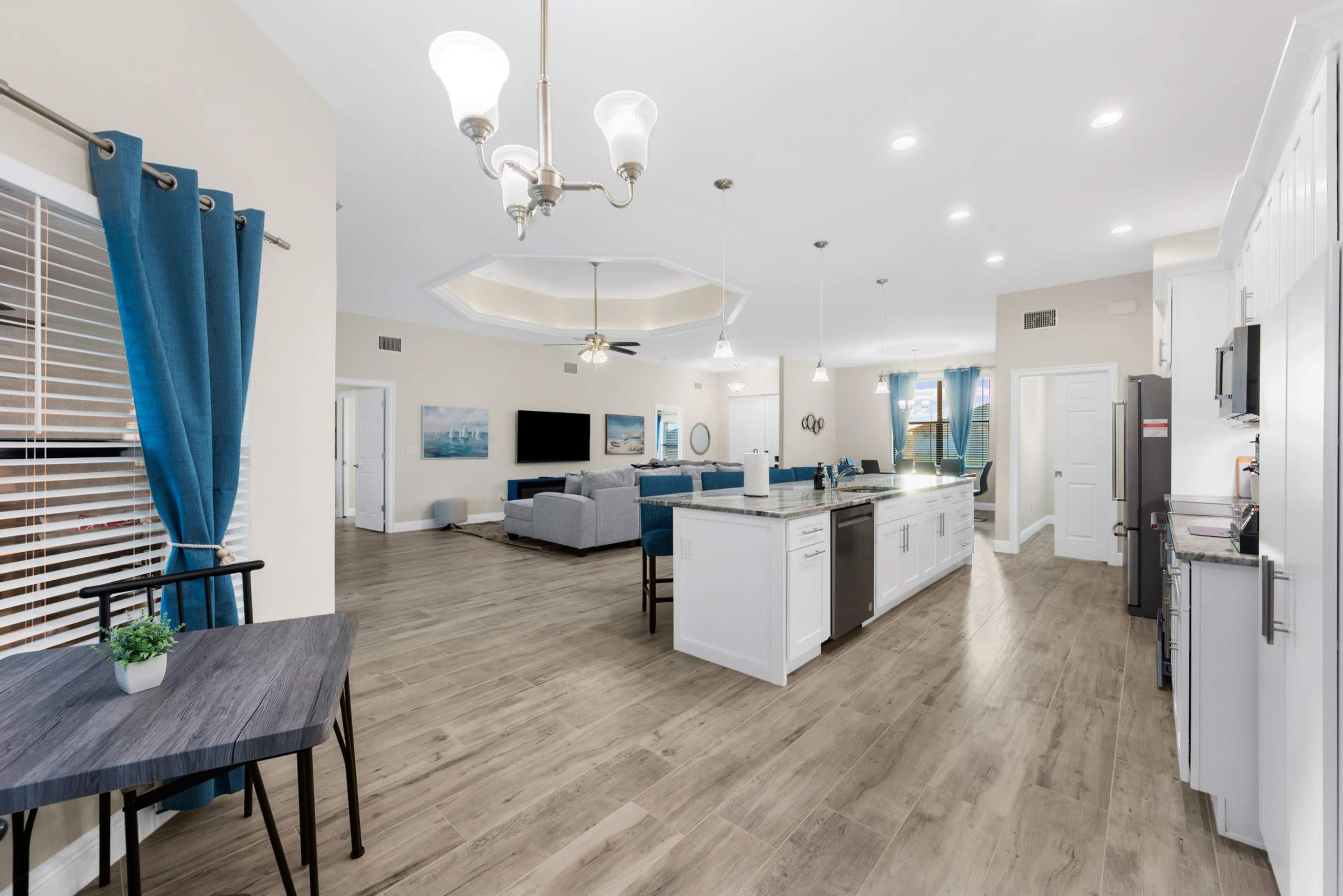 A spacious living area with a kitchen, dining table, gray sofas, and large windows dressed in blue curtains.