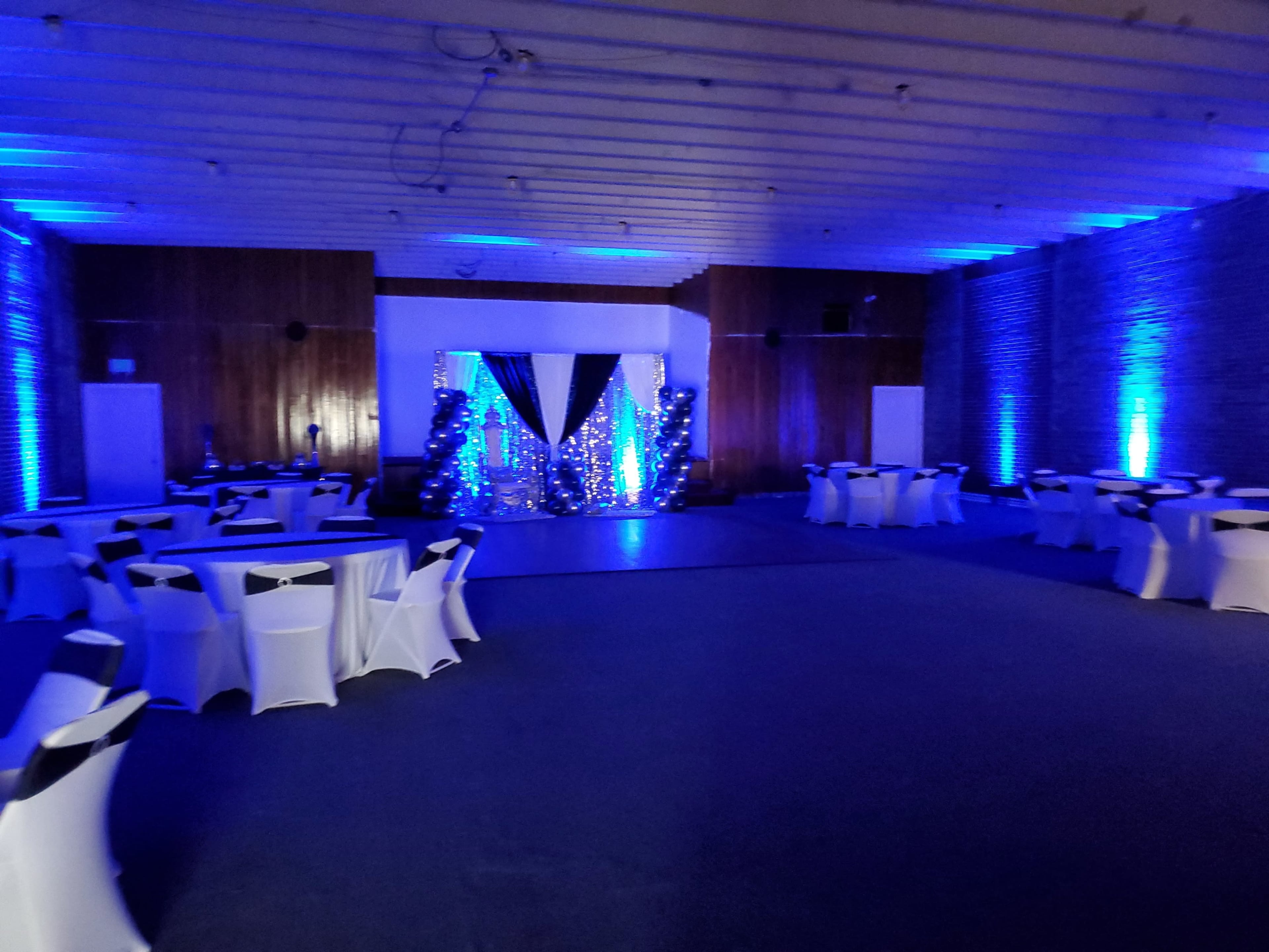 A large event space with tables covered in white cloth, black chair sashes, and a stage adorned with blue and silver decorations at the front.
