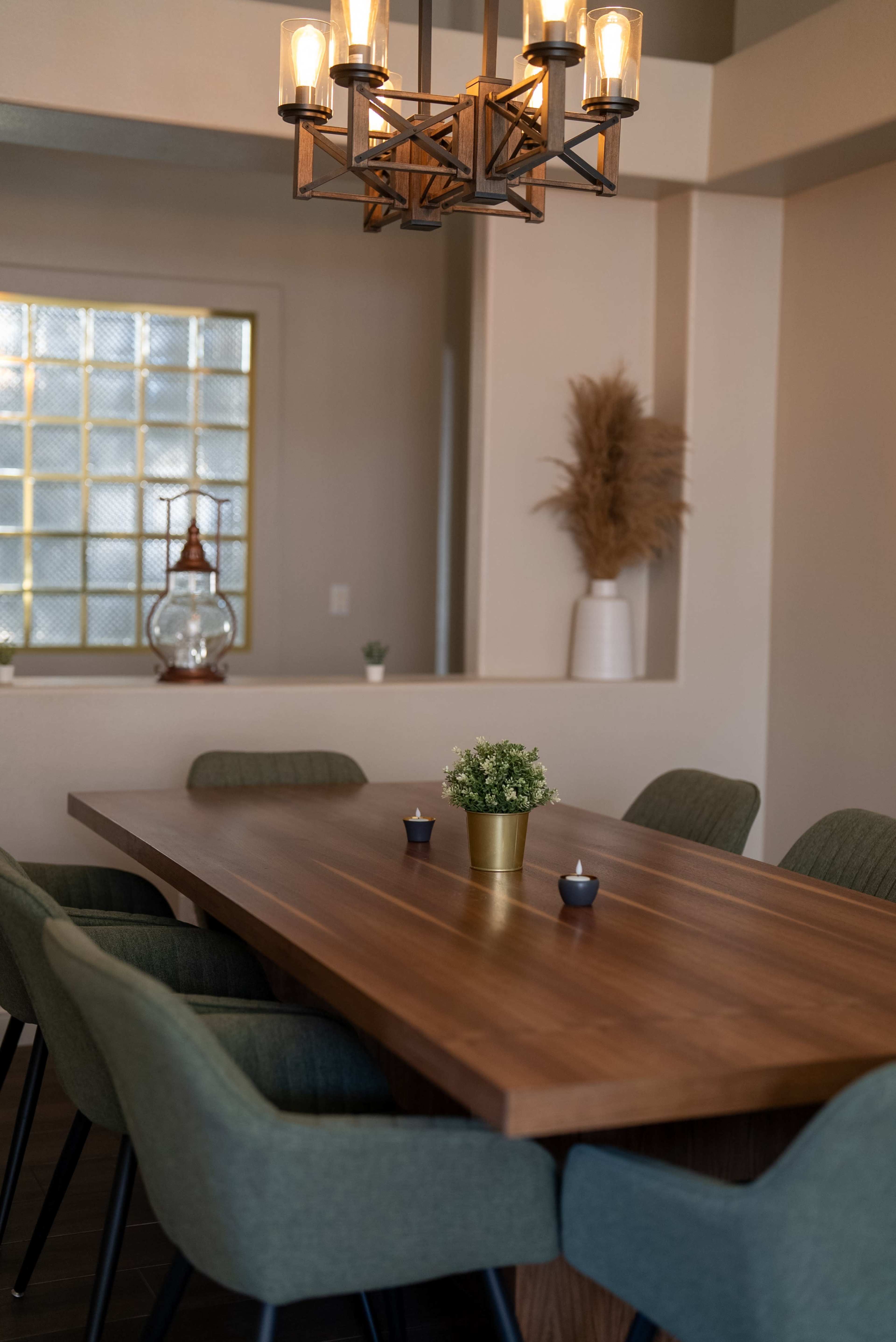 A wooden dining table is surrounded by upholstered chairs, with a chandelier hanging above and decorative plants in the background.