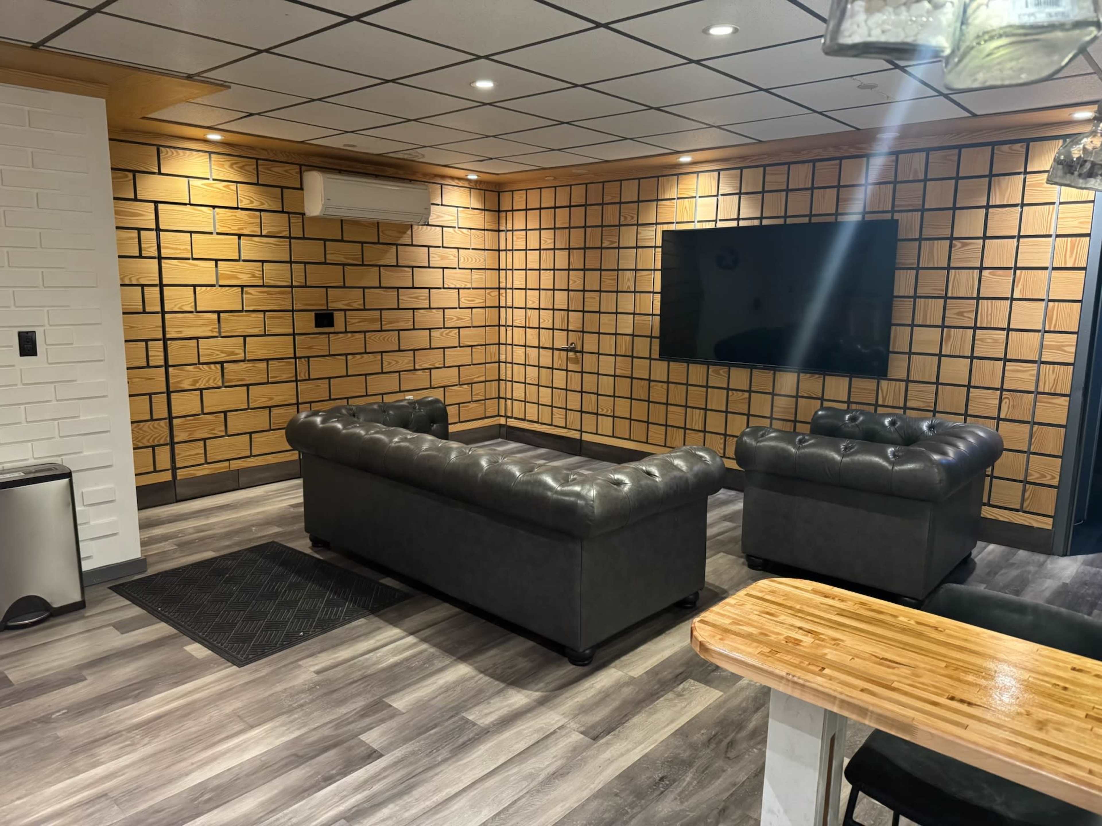 The image shows a modern living room with two dark leather couches, a large flat-screen TV mounted on a wooden-patterned wall, and wooden flooring.