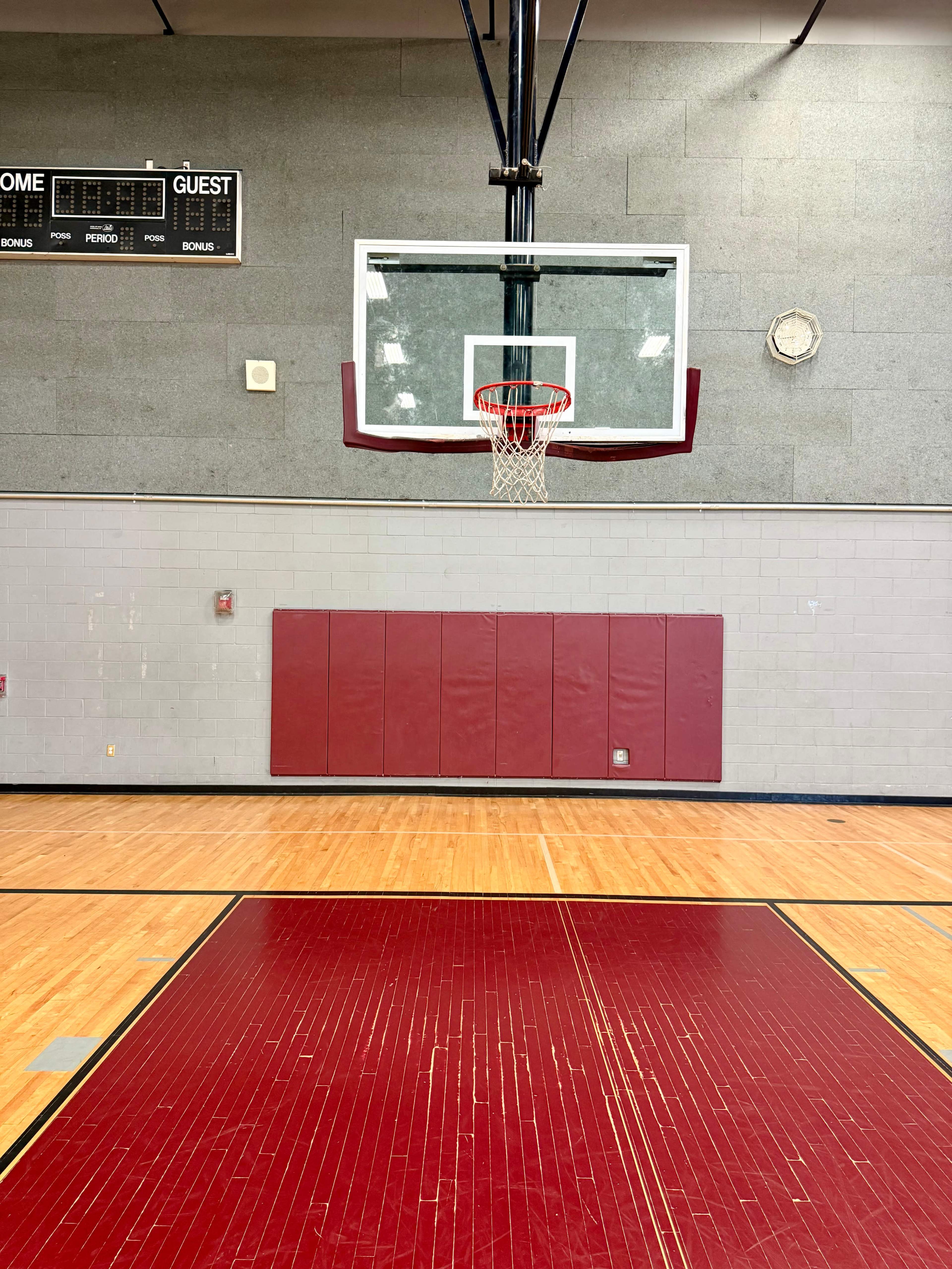 The image shows a basketball hoop and backboard in a gymnasium with wooden flooring and a scoreboard in the background.