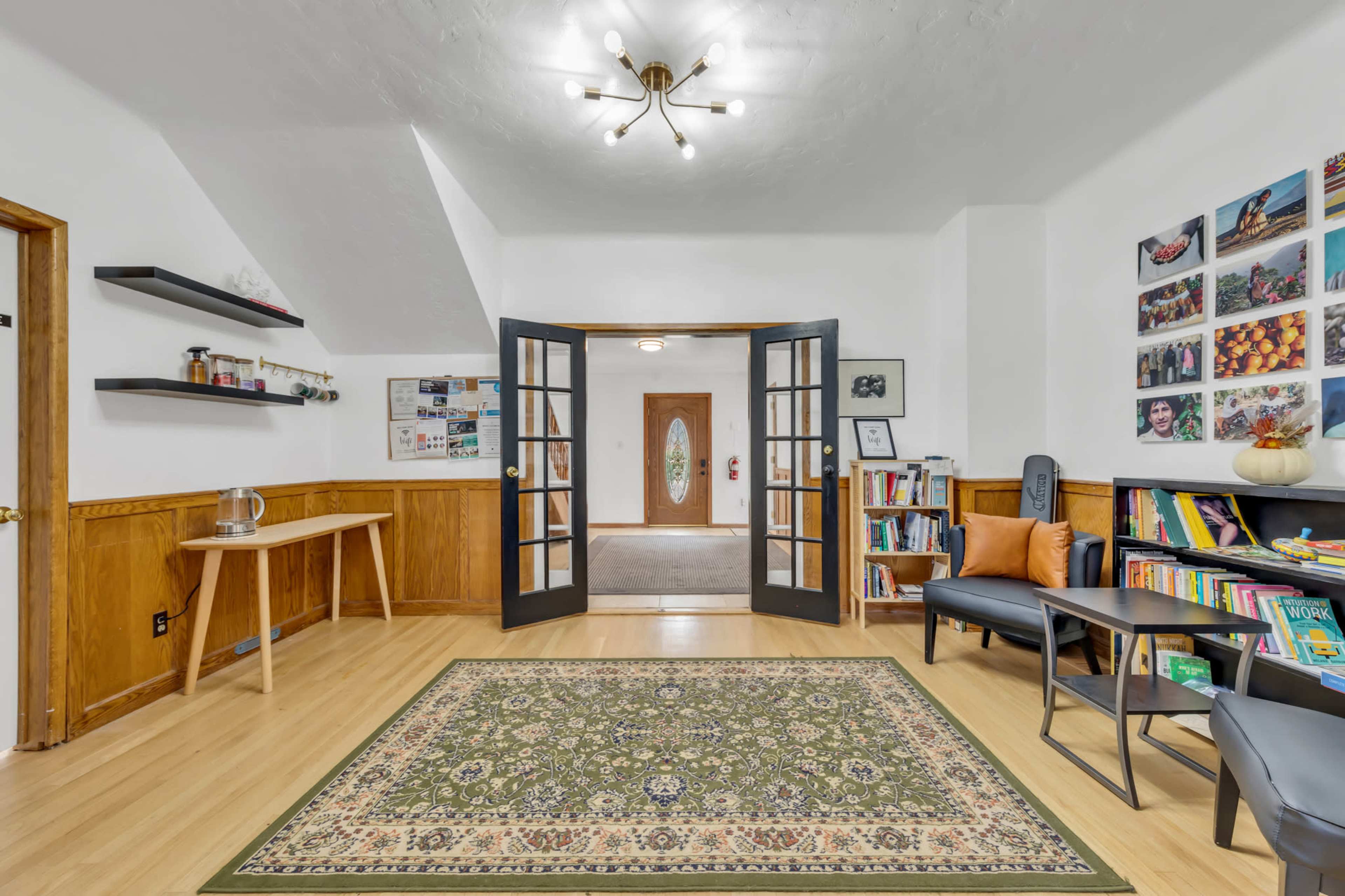 A spacious room with wooden accents, featuring a patterned area rug, a small table, two chairs, and shelves displaying books and decor.