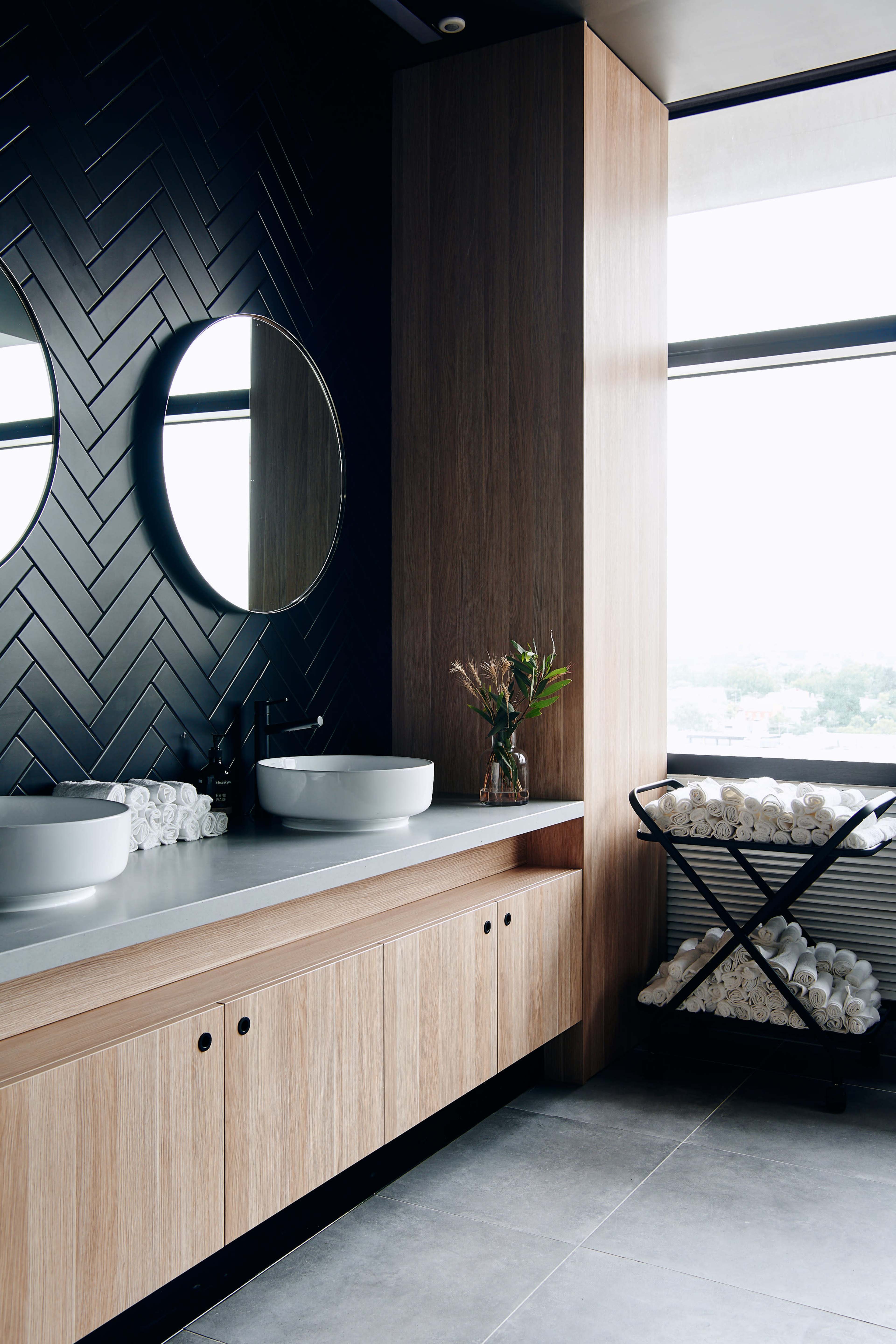 The bathroom features two circular mirrors above vessel sinks, a wooden vanity with storage, and a cart stacked with rolled towels next to a large window.