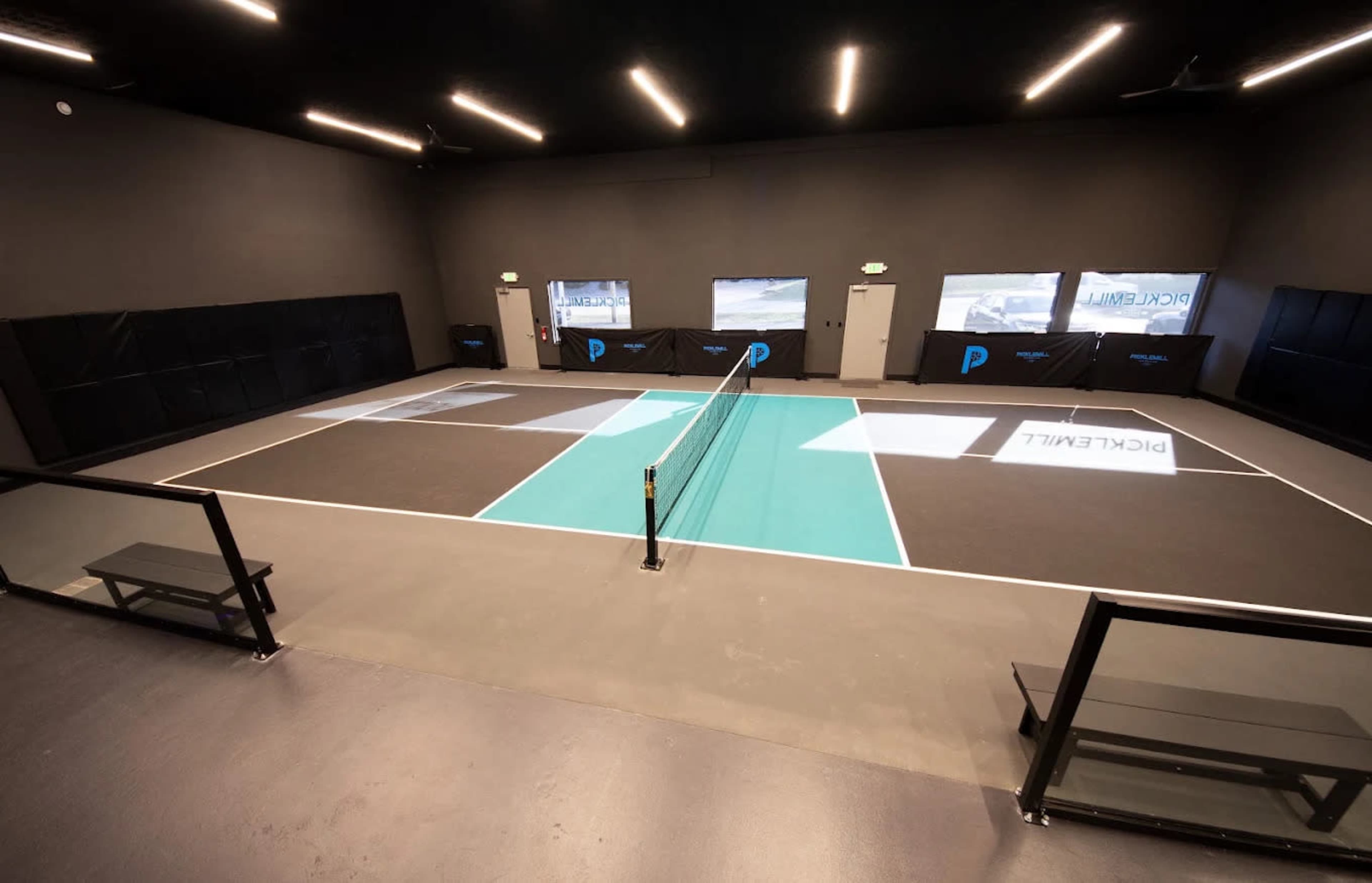 The image shows an indoor pickleball court with a teal-colored playing area, surrounded by black walls and large windows.