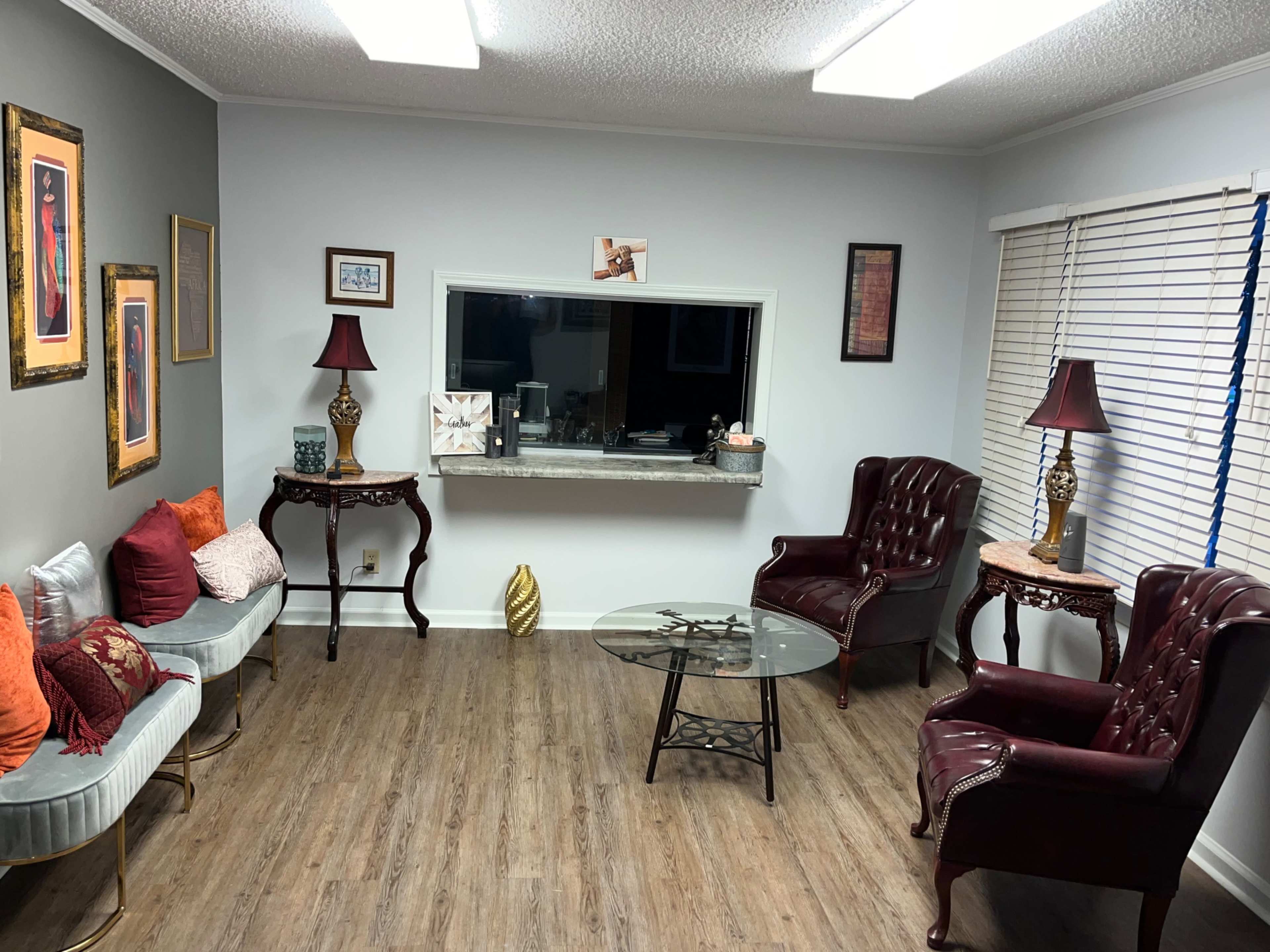 The image depicts a cozy living room with two maroon armchairs, a glass coffee table, and a mixture of decorative items and artwork on the walls.