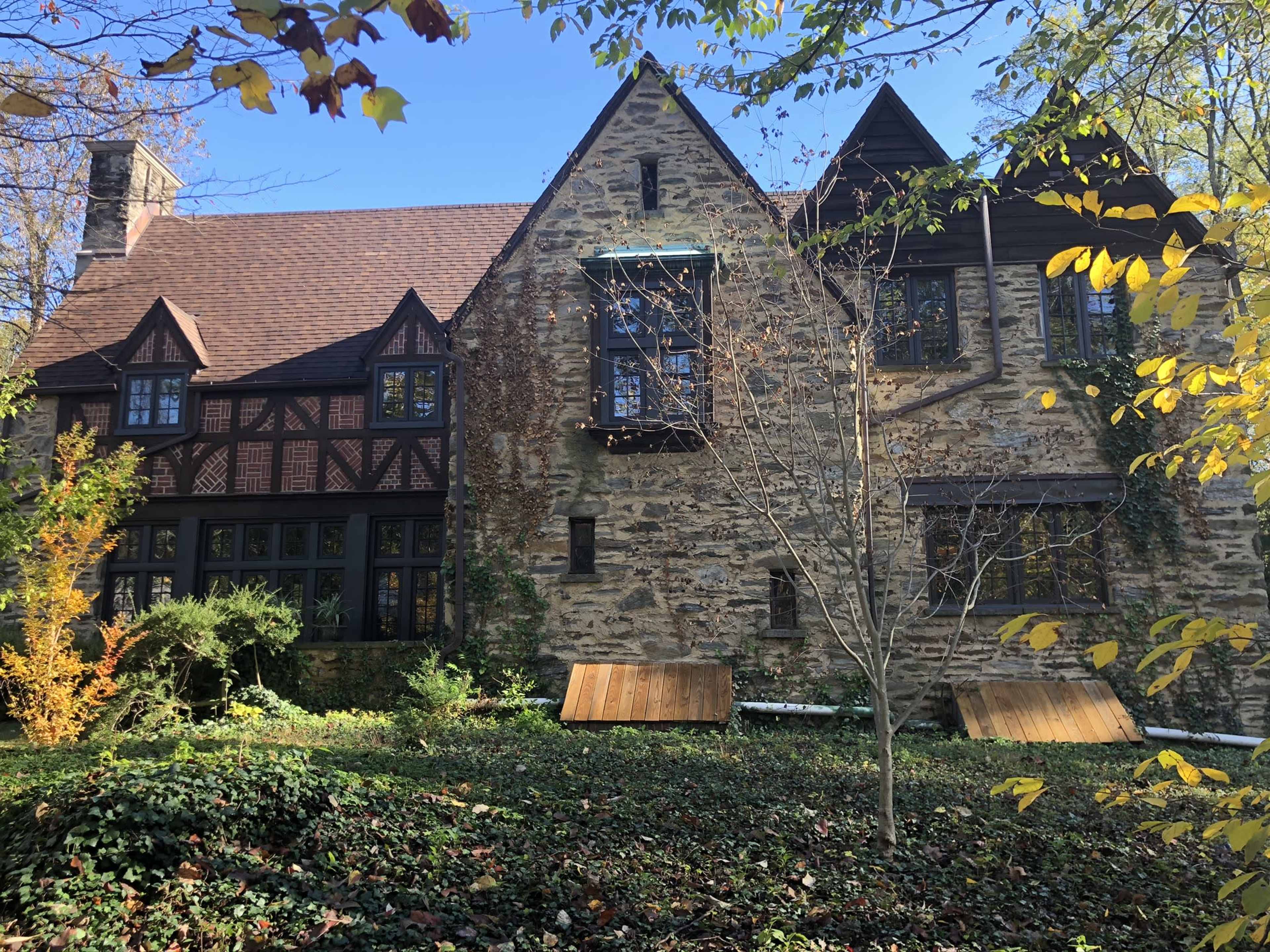 A stone and wood house features a mix of architectural styles with large windows and a sloped roof, surrounded by greenery and autumn foliage.