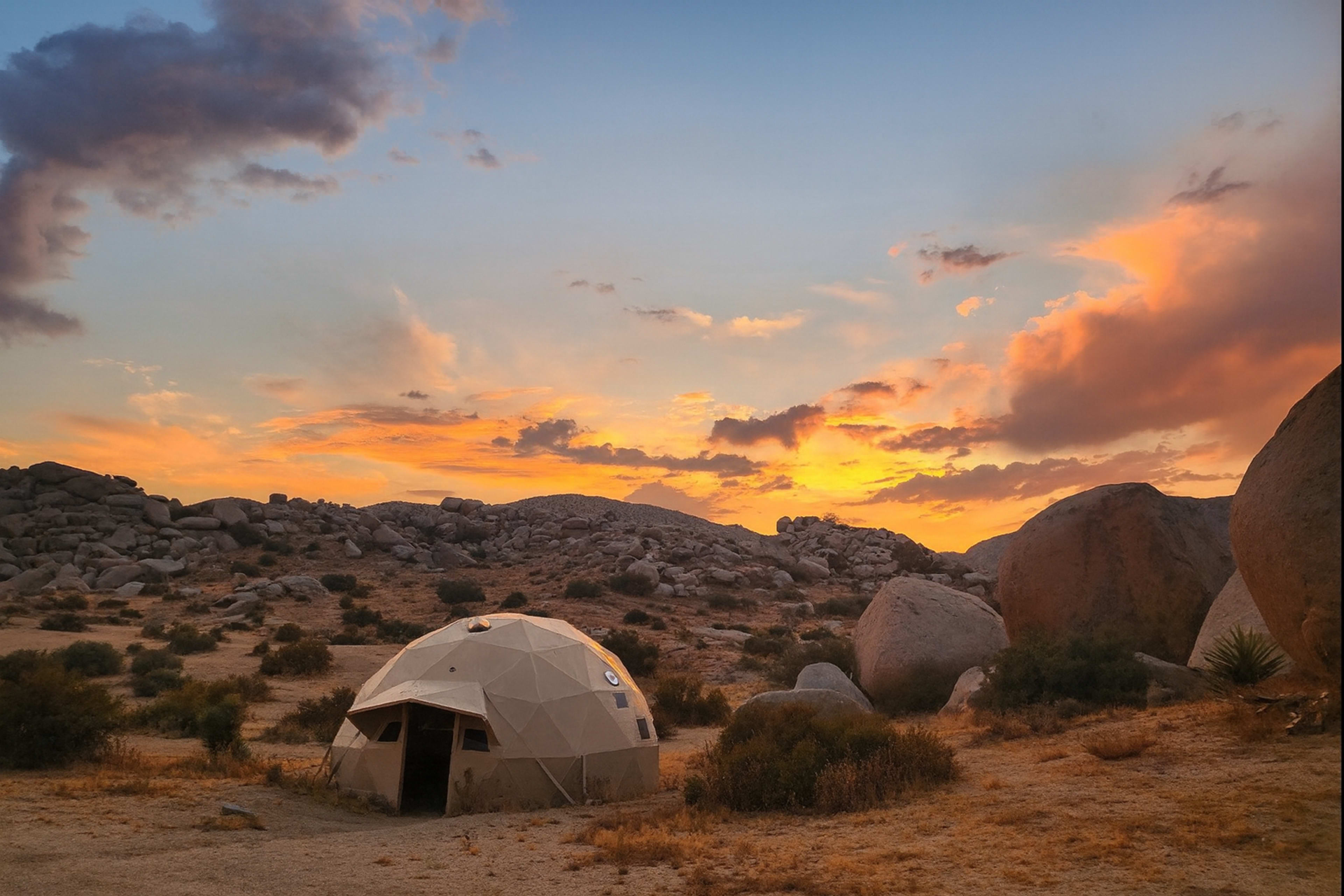A geodesic dome sits in a rocky desert landscape under a colorful sunset sky.