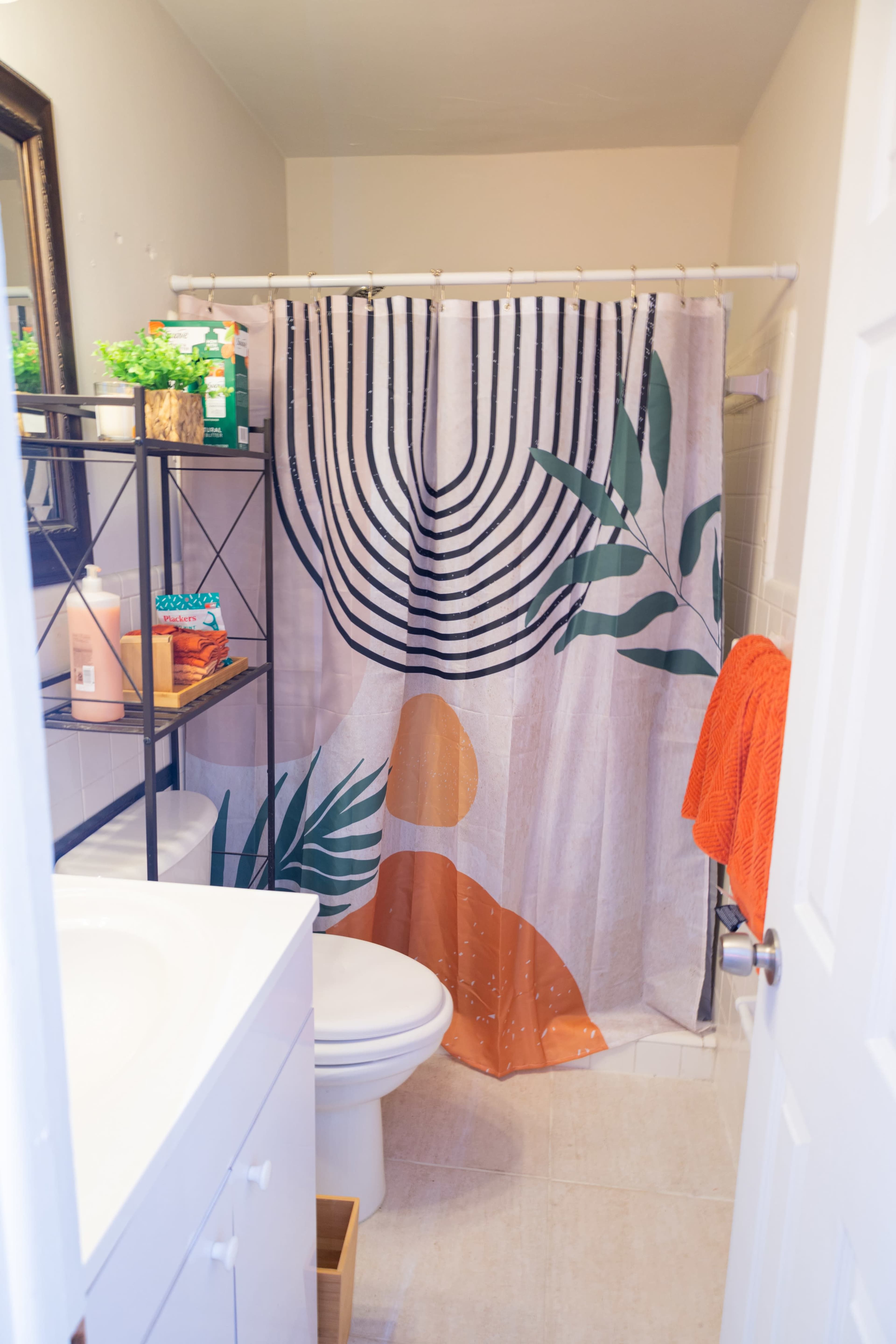 A bathroom features a shower with a decorative curtain displaying geometric patterns and plant motifs, along with a white vanity and toiletries on a shelf.