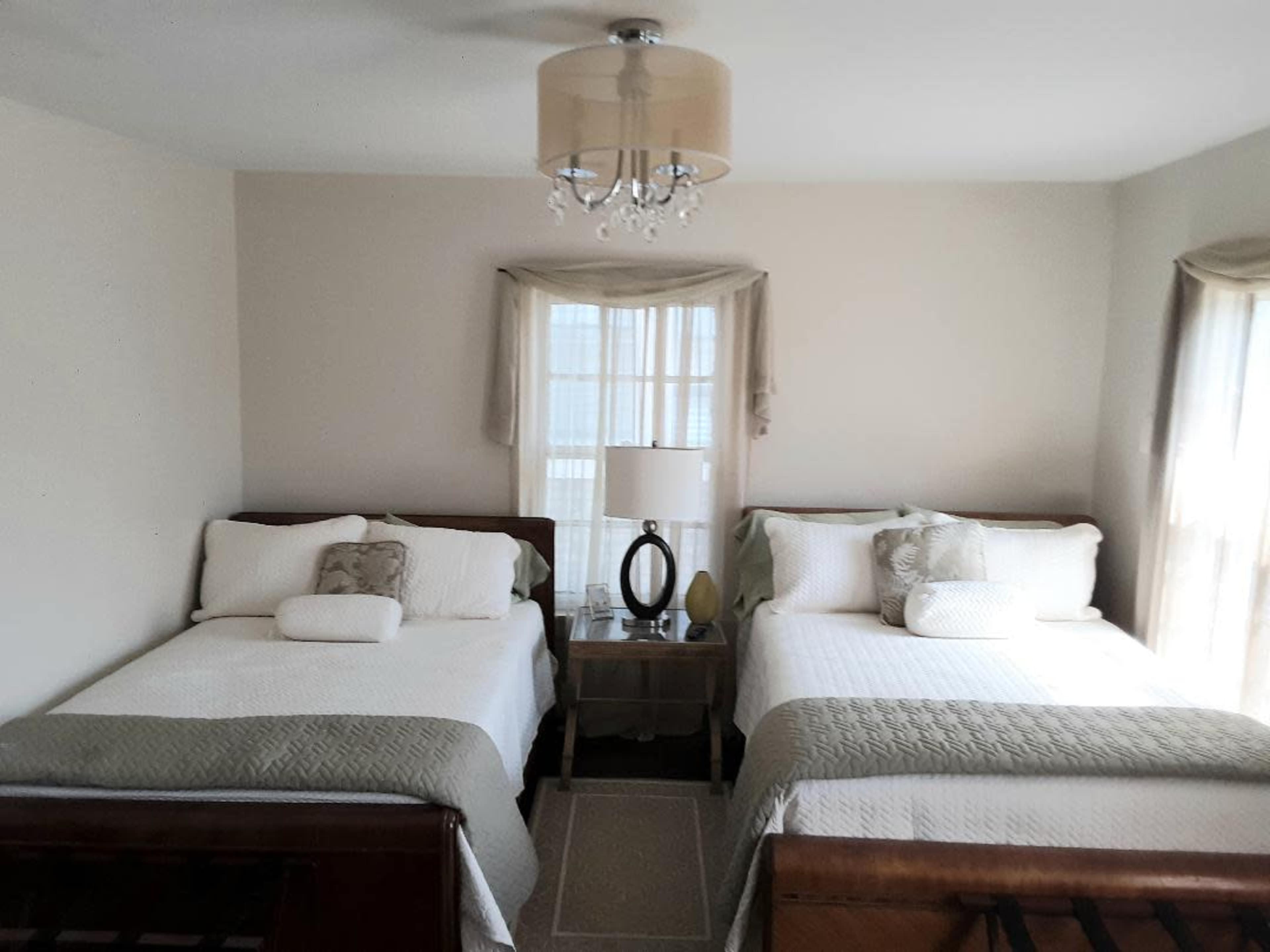 A room with two beds, each with white bedding and decorative pillows, a lamp on a nightstand, and large windows with sheer curtains.