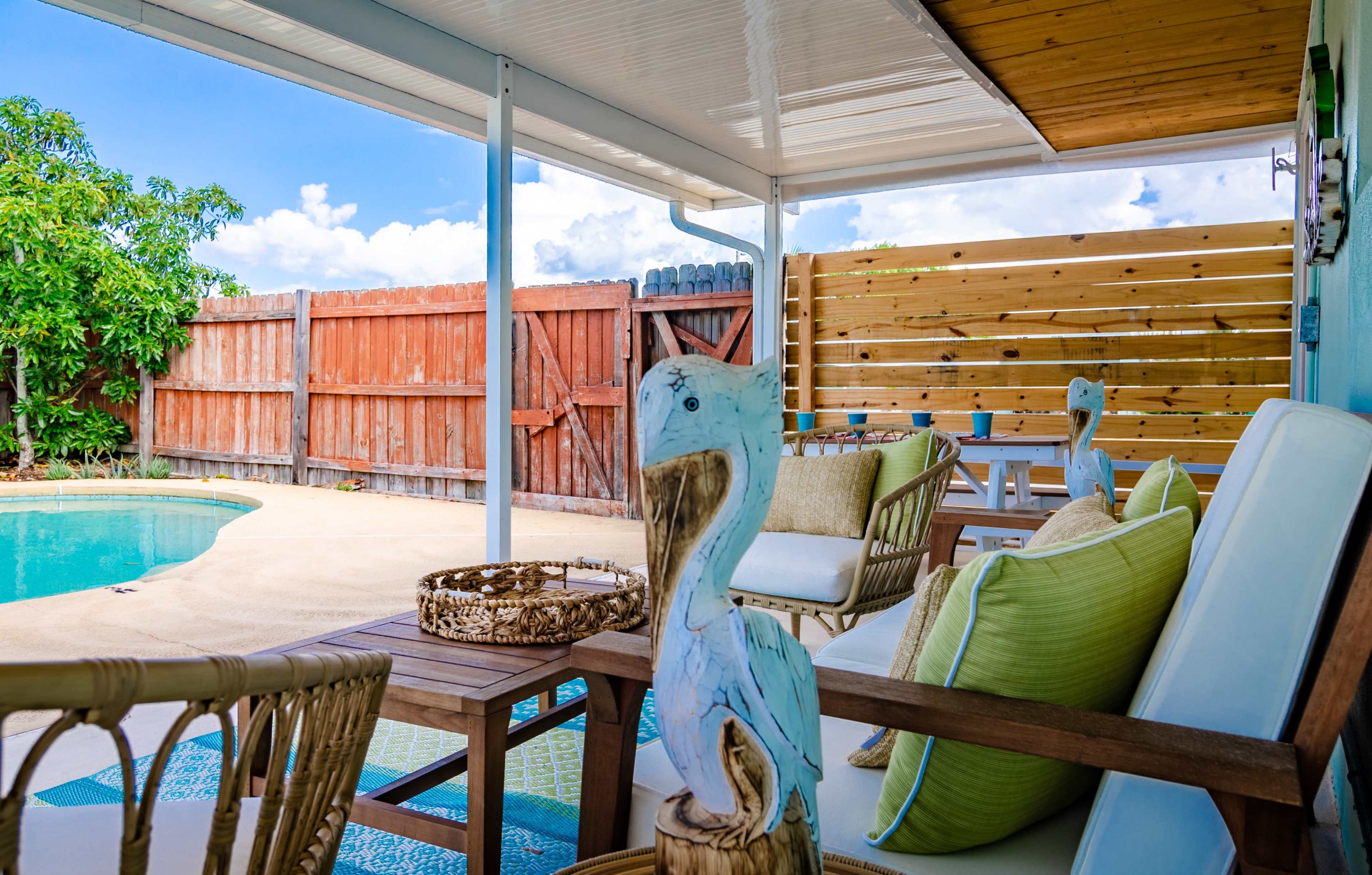 A shaded patio with wicker chairs and decorative pelican figures overlooks a swimming pool surrounded by a wooden fence.
