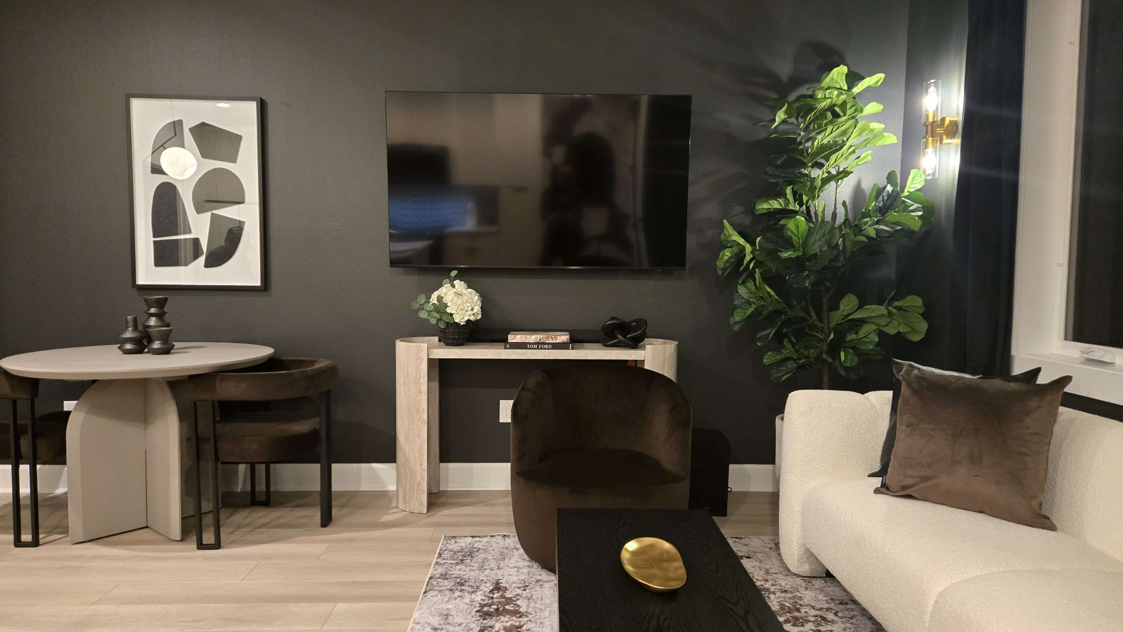 The image shows a modern living room featuring a television mounted on a wall, a round dining table with chairs, a light-colored couch, and decorative plants.