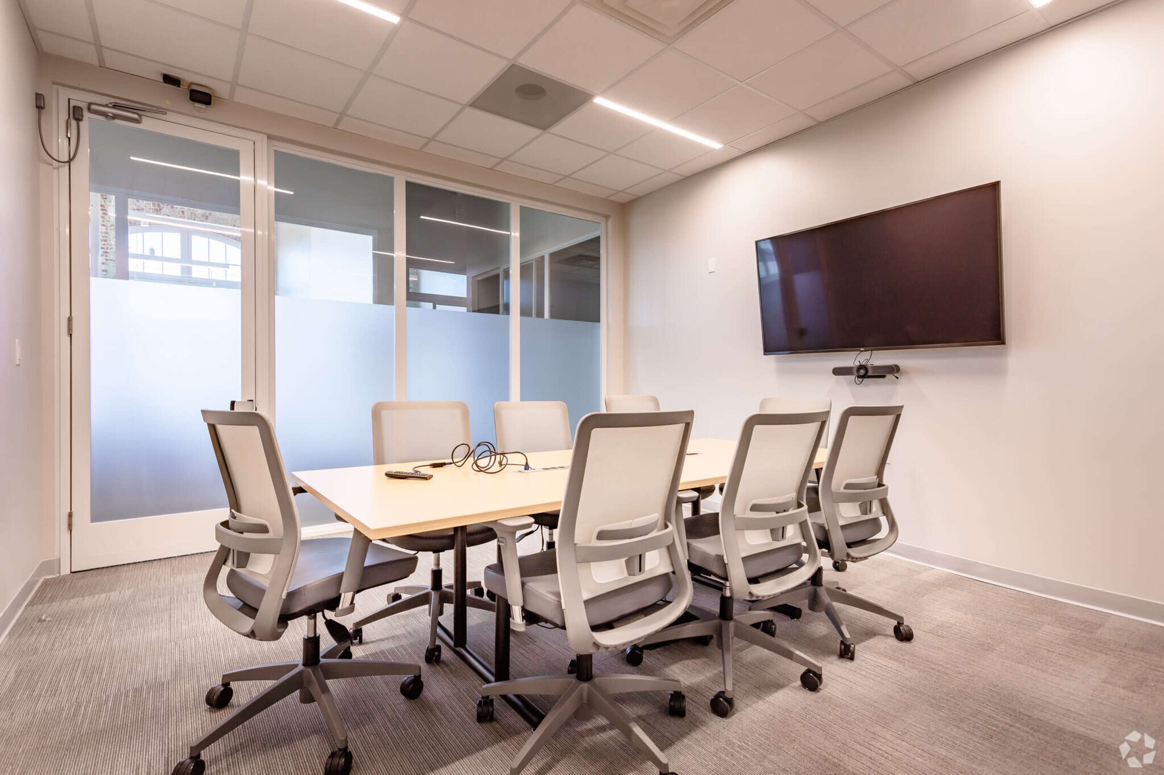 A modern conference room features a long table surrounded by six ergonomic chairs, with a wall-mounted TV and glass-paneled doors.