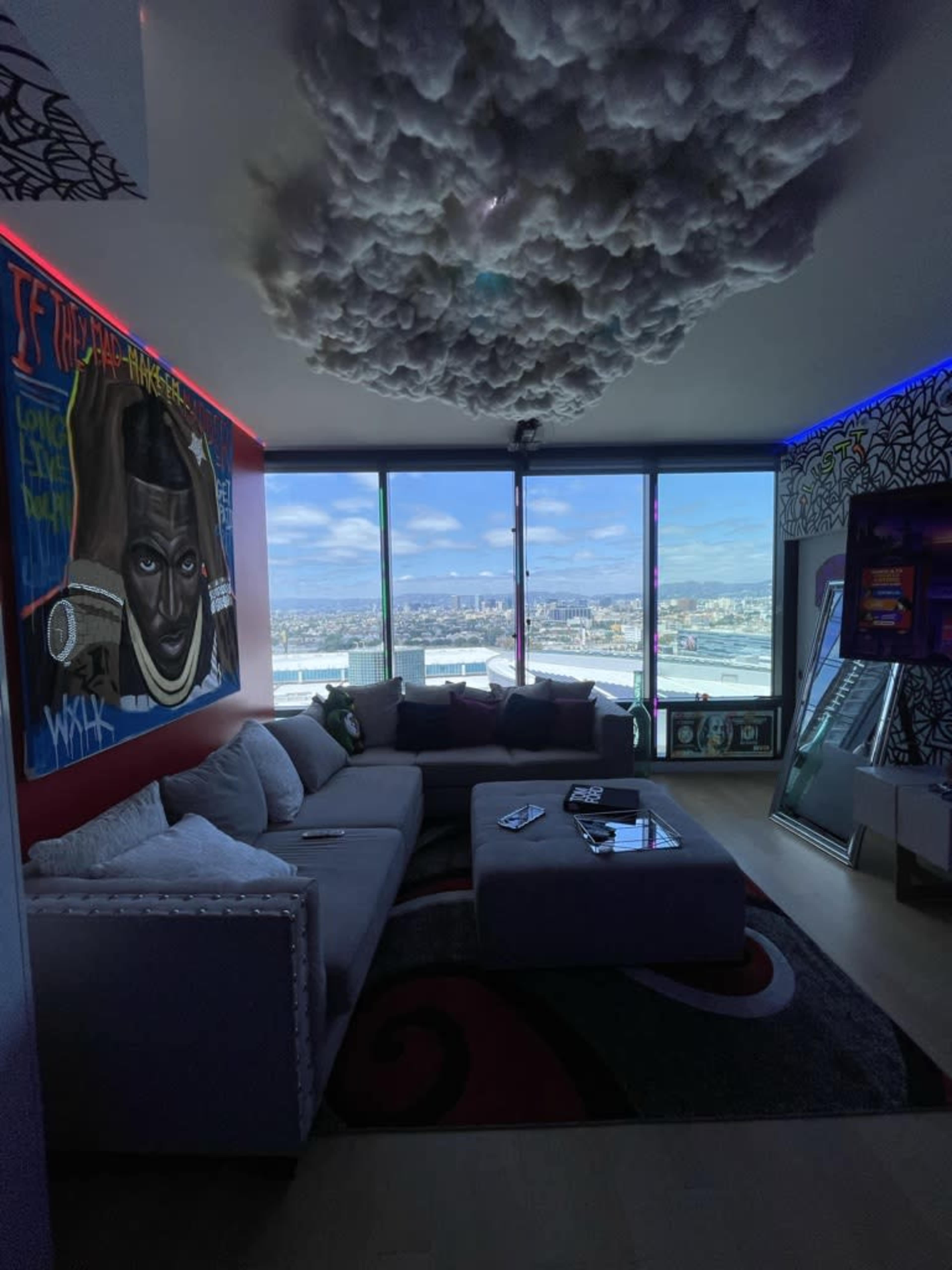 The image shows a modern living room with a gray sectional sofa, a coffee table, and large windows offering a view of the city skyline, complemented by colorful artwork and decorative lighting.