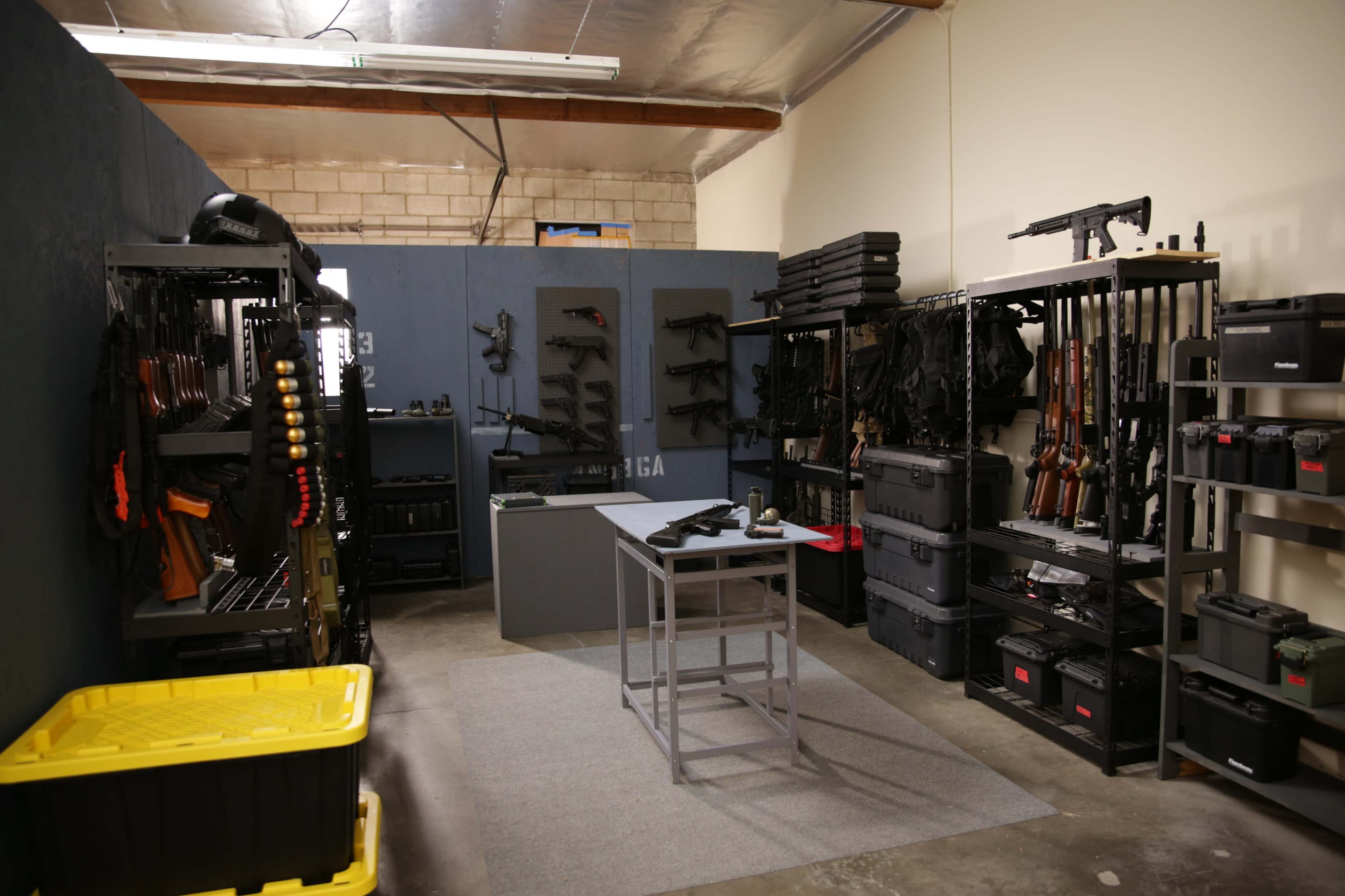 The image shows an organized room filled with various firearms and tactical gear displayed on shelves and racks, with a central table set for inspection or maintenance.