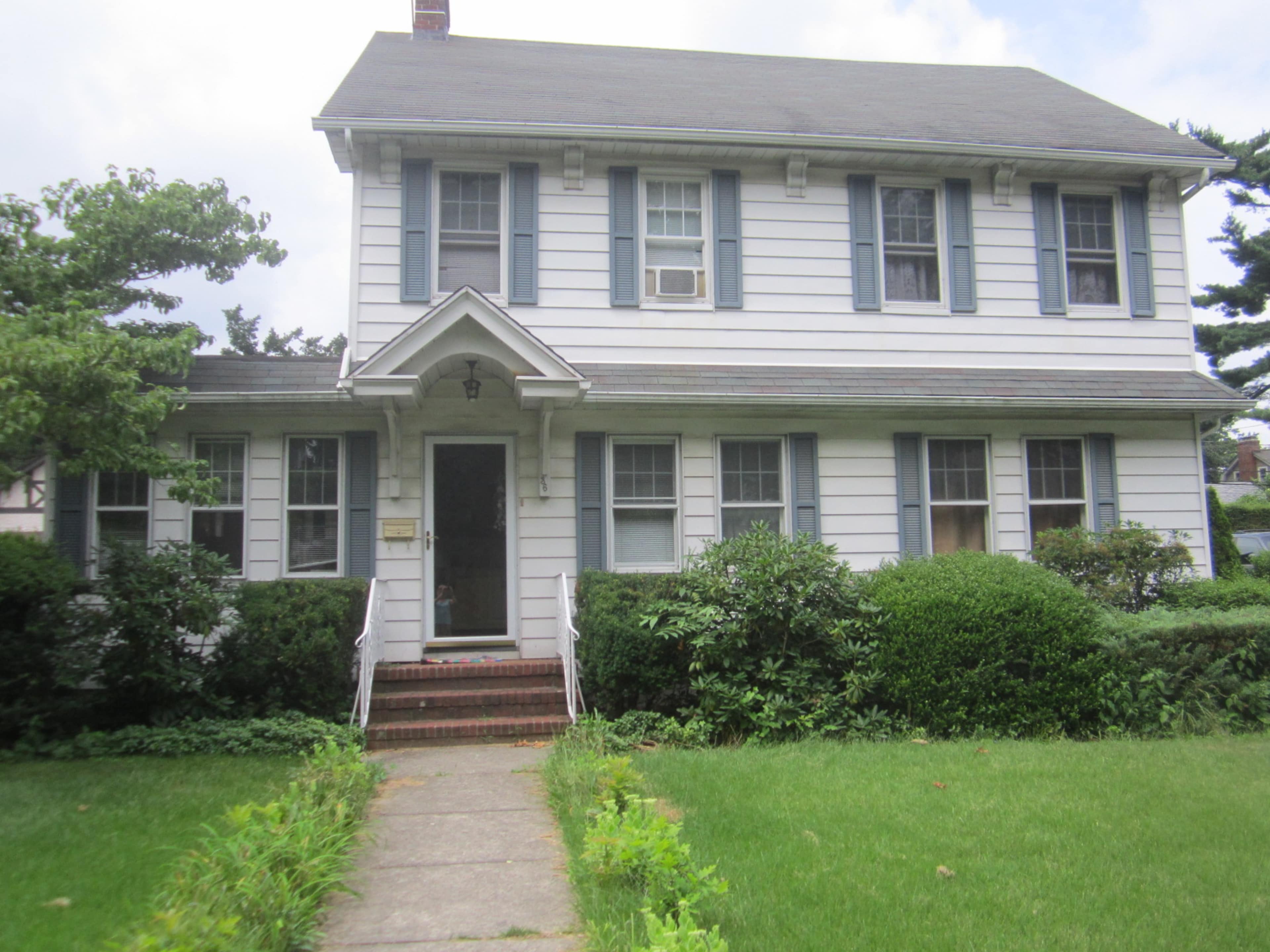 A two-story, white wooden house with dark blue shutters and a small front porch surrounded by green shrubs and a lawn.