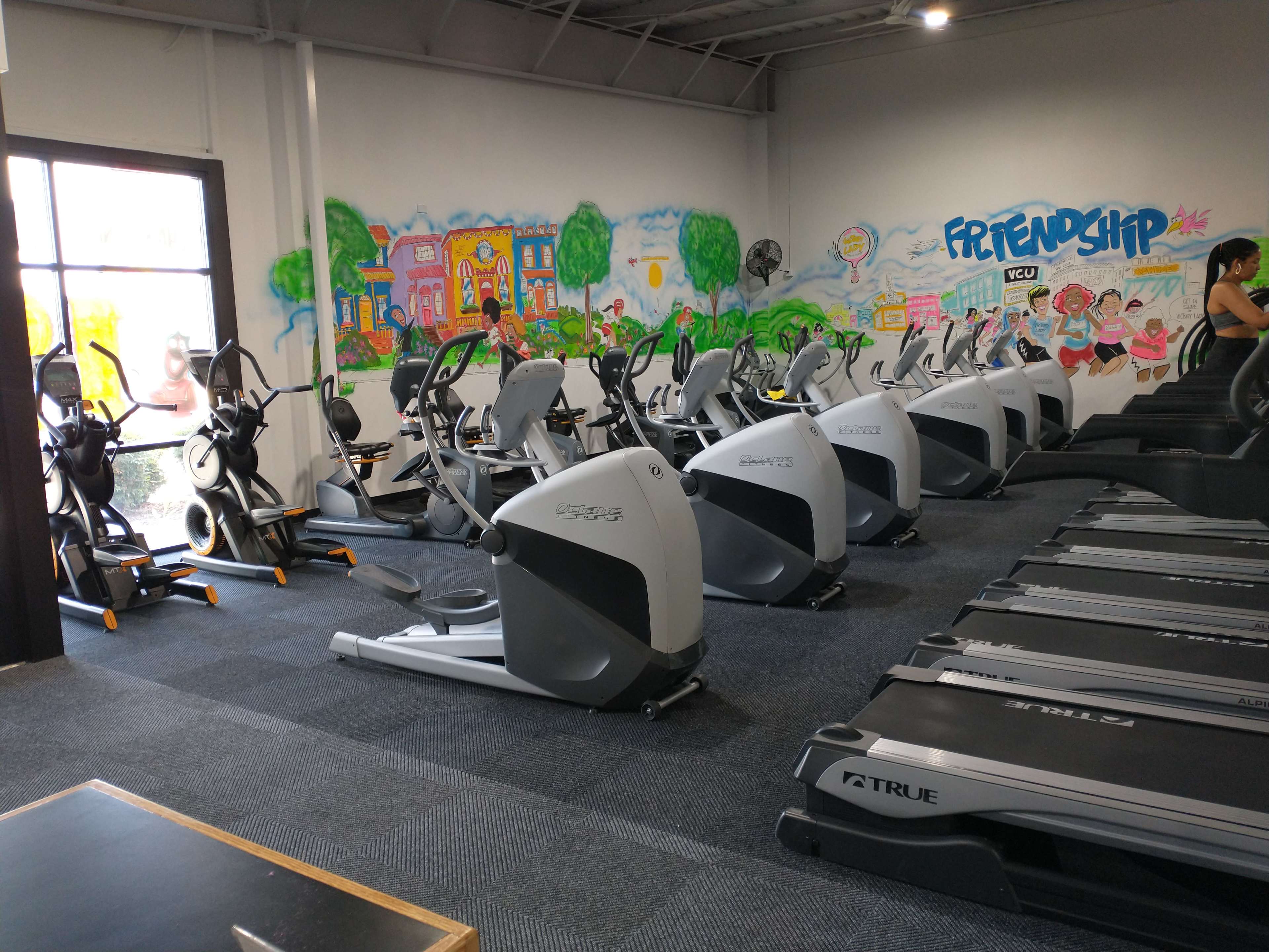 A gym interior with several elliptical machines and treadmills arranged in a fitness space featuring a colorful mural on the wall.