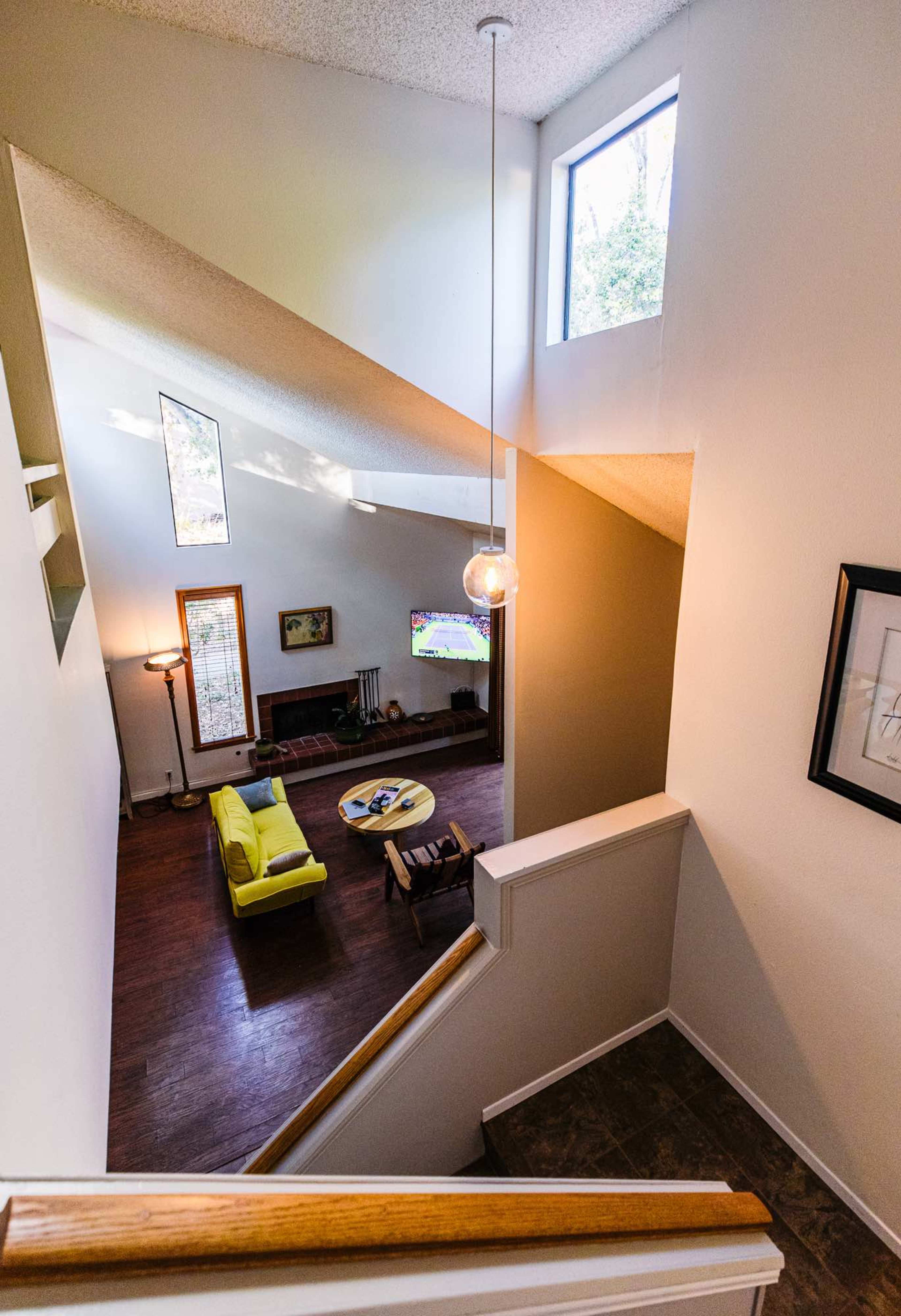 A staircase leads down to a living room with a yellow sofa and coffee table, illuminated by natural light from windows.