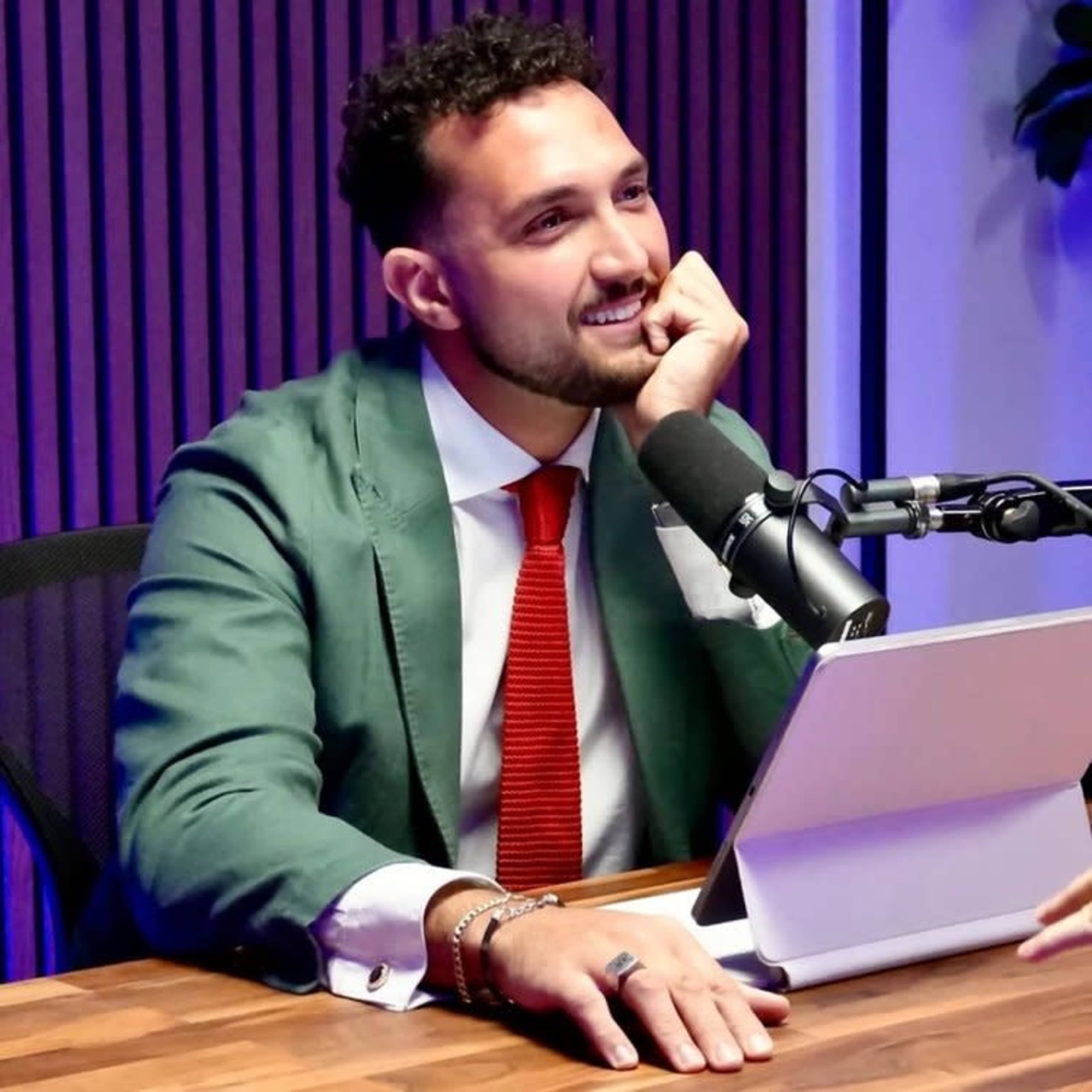 A man in a green suit and red tie sits at a desk with a tablet and microphone in front of him, smiling while engaged in a conversation.