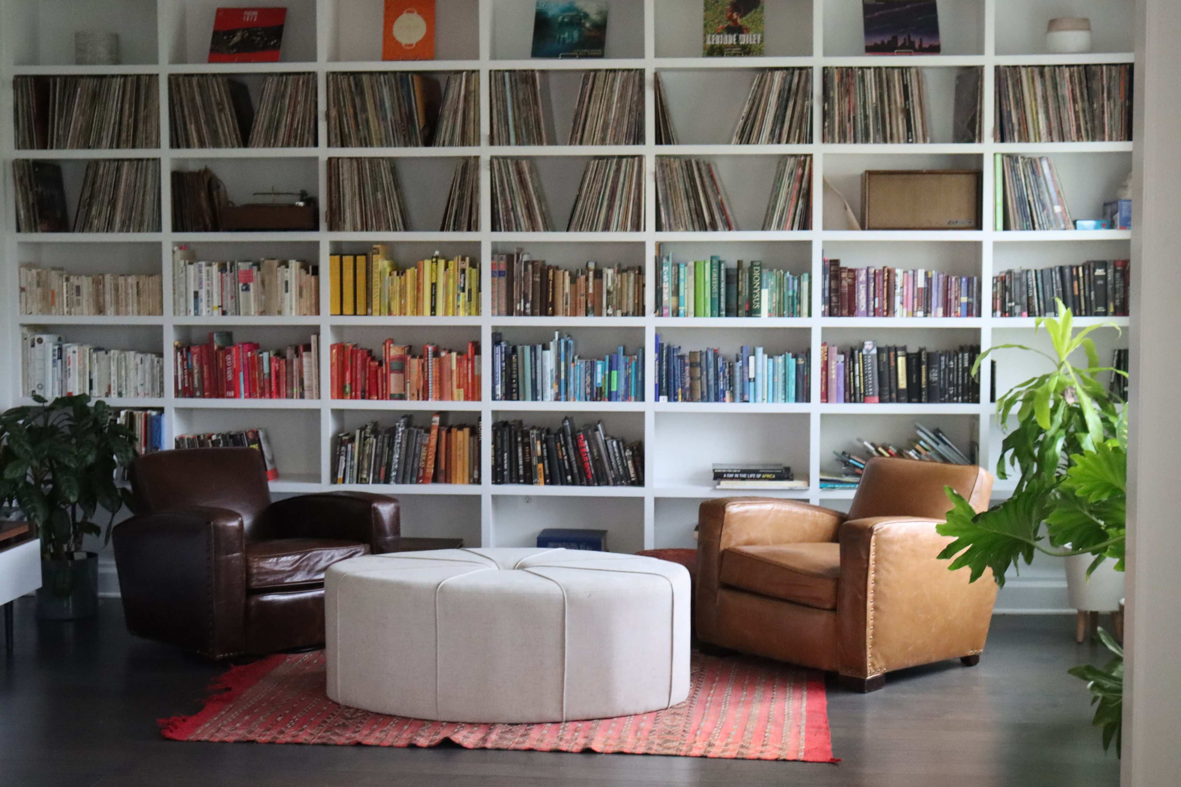 A cozy reading nook features two leather armchairs facing a circular ottoman, surrounded by a wall of books organized by color.