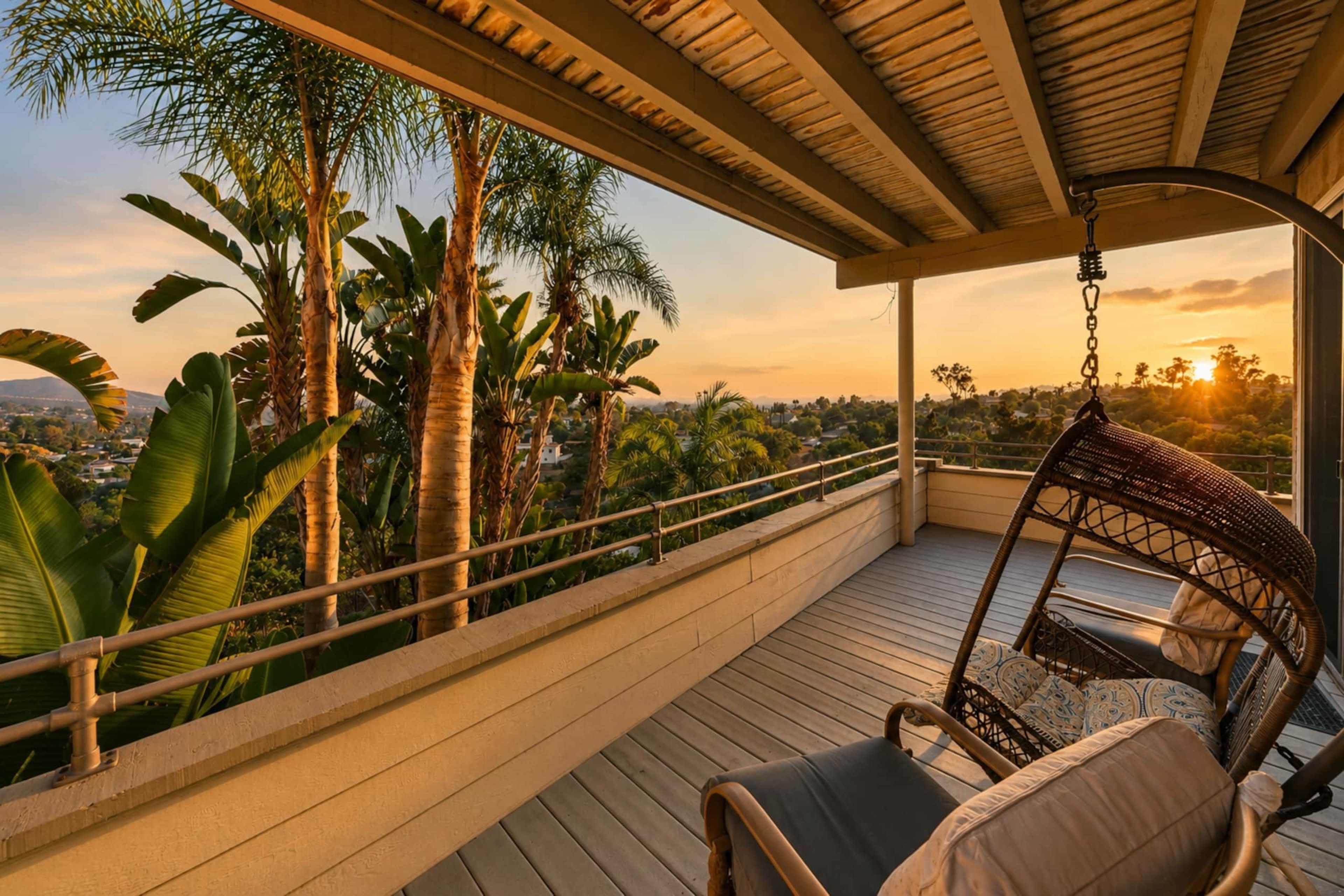 The image shows a balcony with a hanging chair, surrounded by tropical plants, overlooking a sunset-lit landscape.
