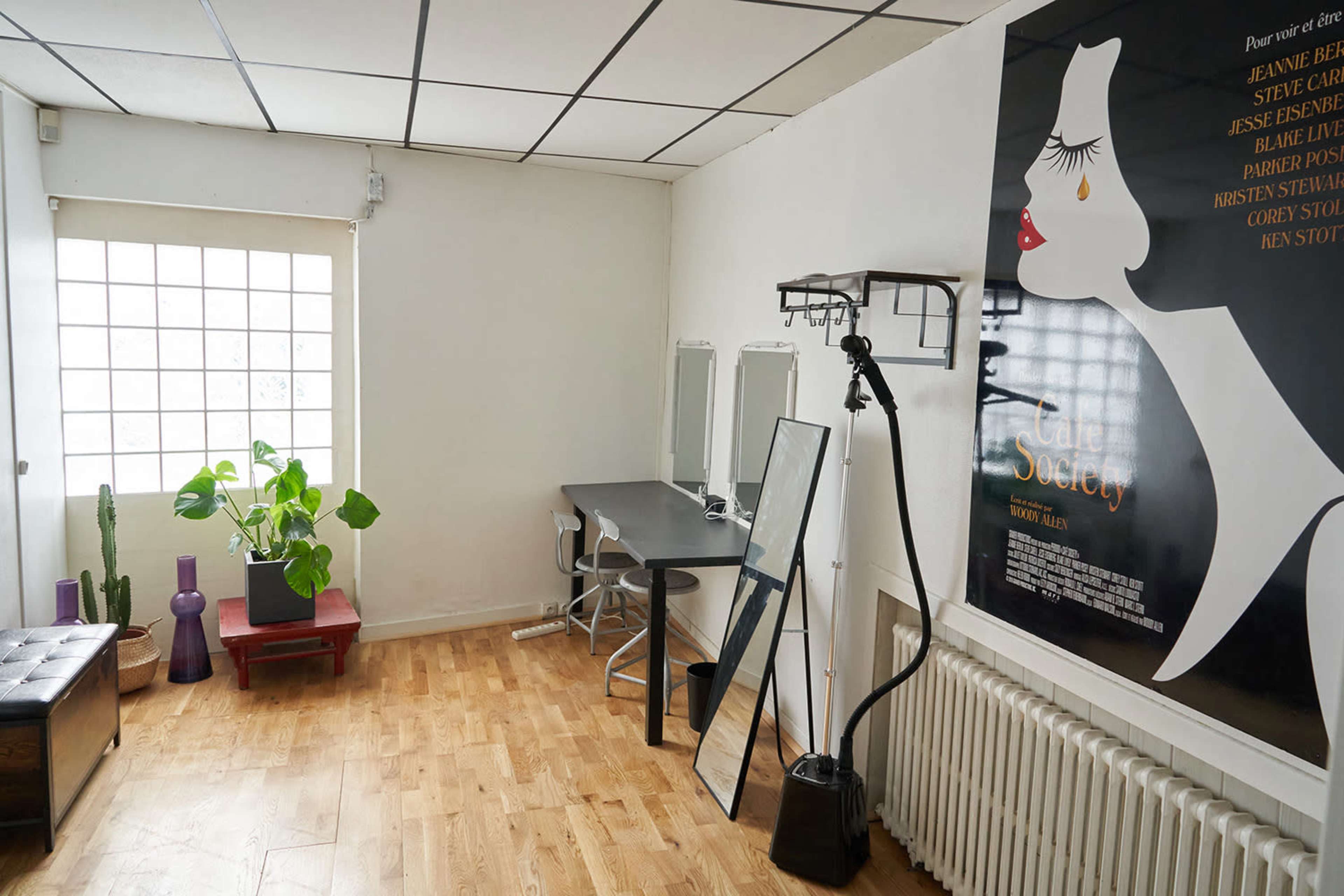 A bright room featuring a desk with a chair, a large mirror, a potted plant, and a film poster on the wall.