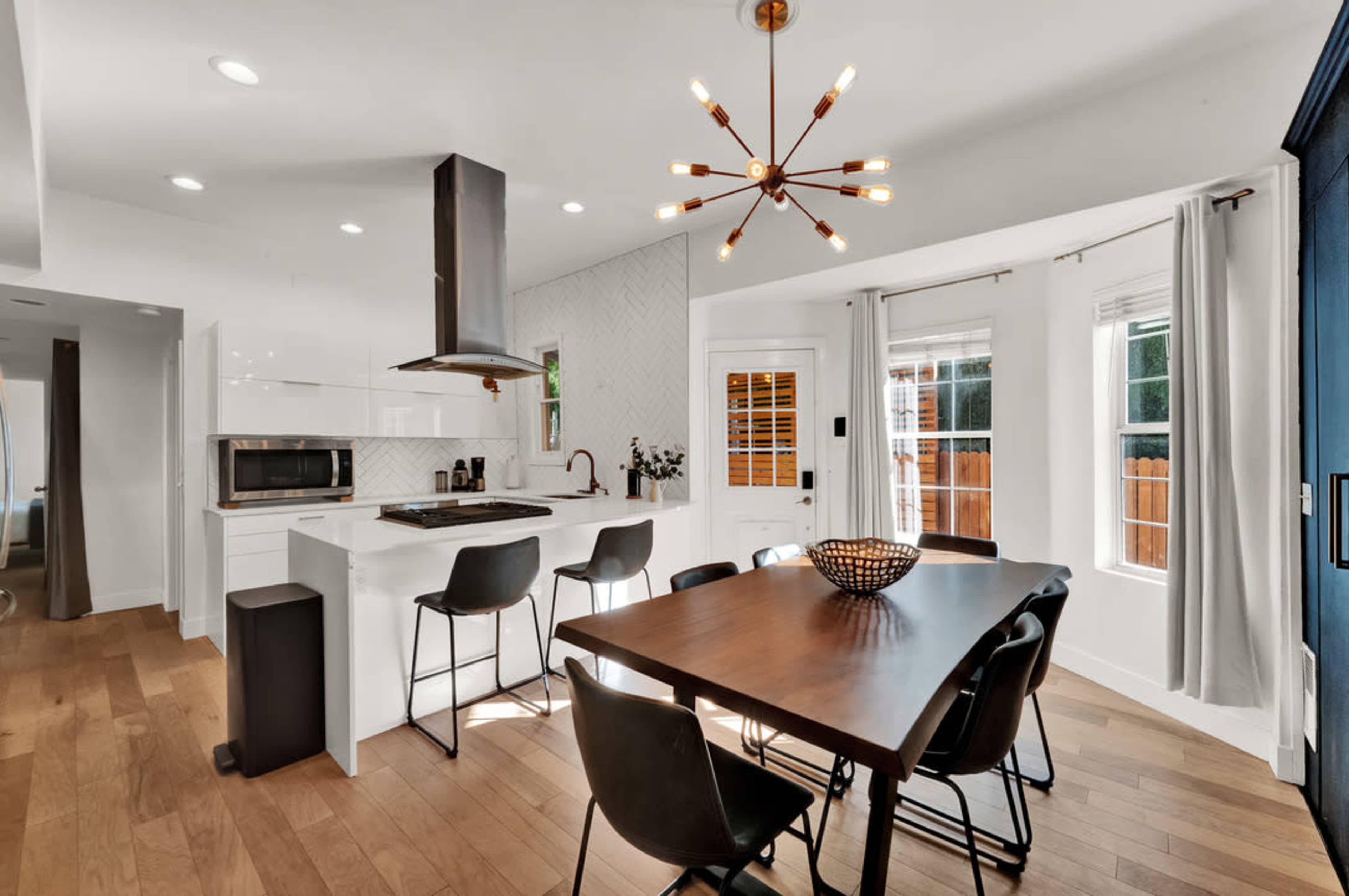 A modern kitchen and dining area featuring a central table with eight black chairs, a white countertop with a stovetop, and a stylish light fixture above.