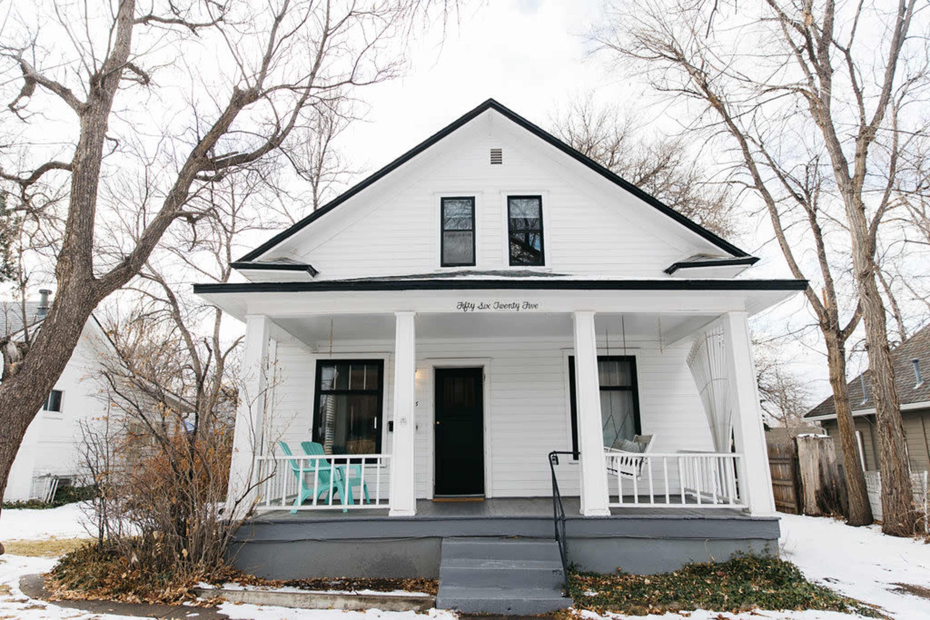 A white two-story house with a covered porch and two turquoise chairs sits among leafless trees on a quiet street.