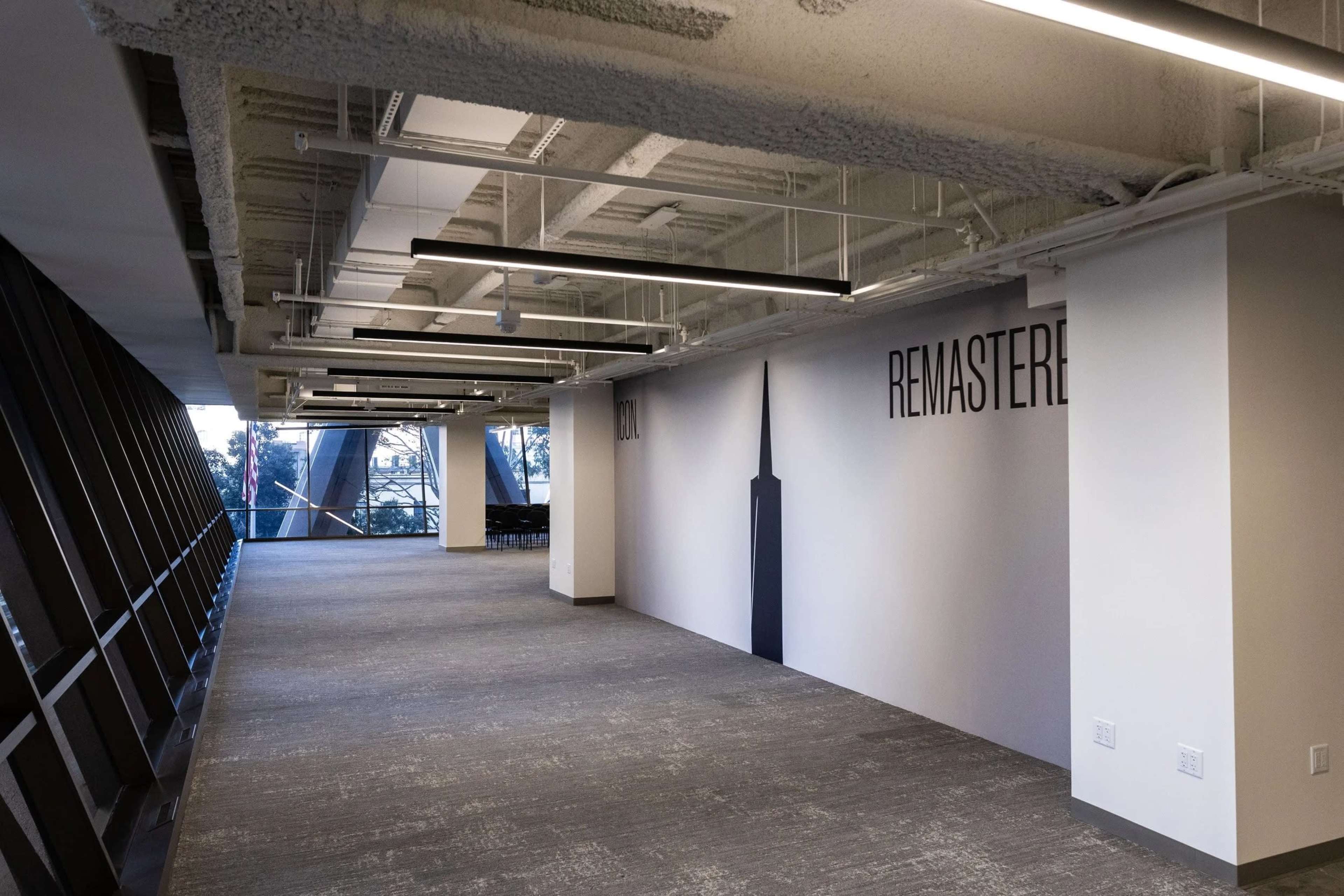 The image shows a modern, empty office corridor with large windows and a wall featuring the word "REMASTERED" painted on it.