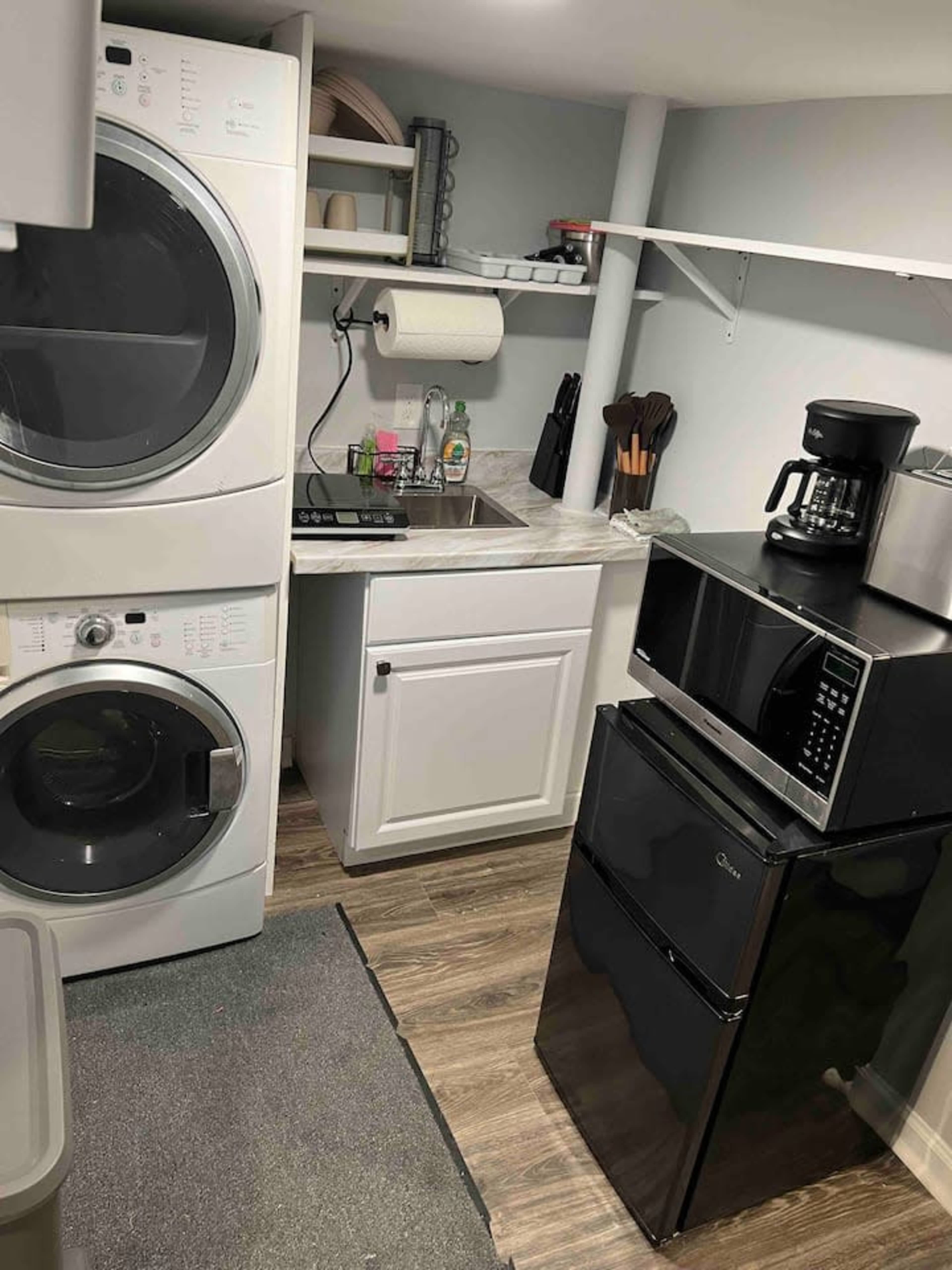 A small laundry area with a stacked washer and dryer, a countertop workspace with a sink, and a microwave on top of a black refrigerator.
