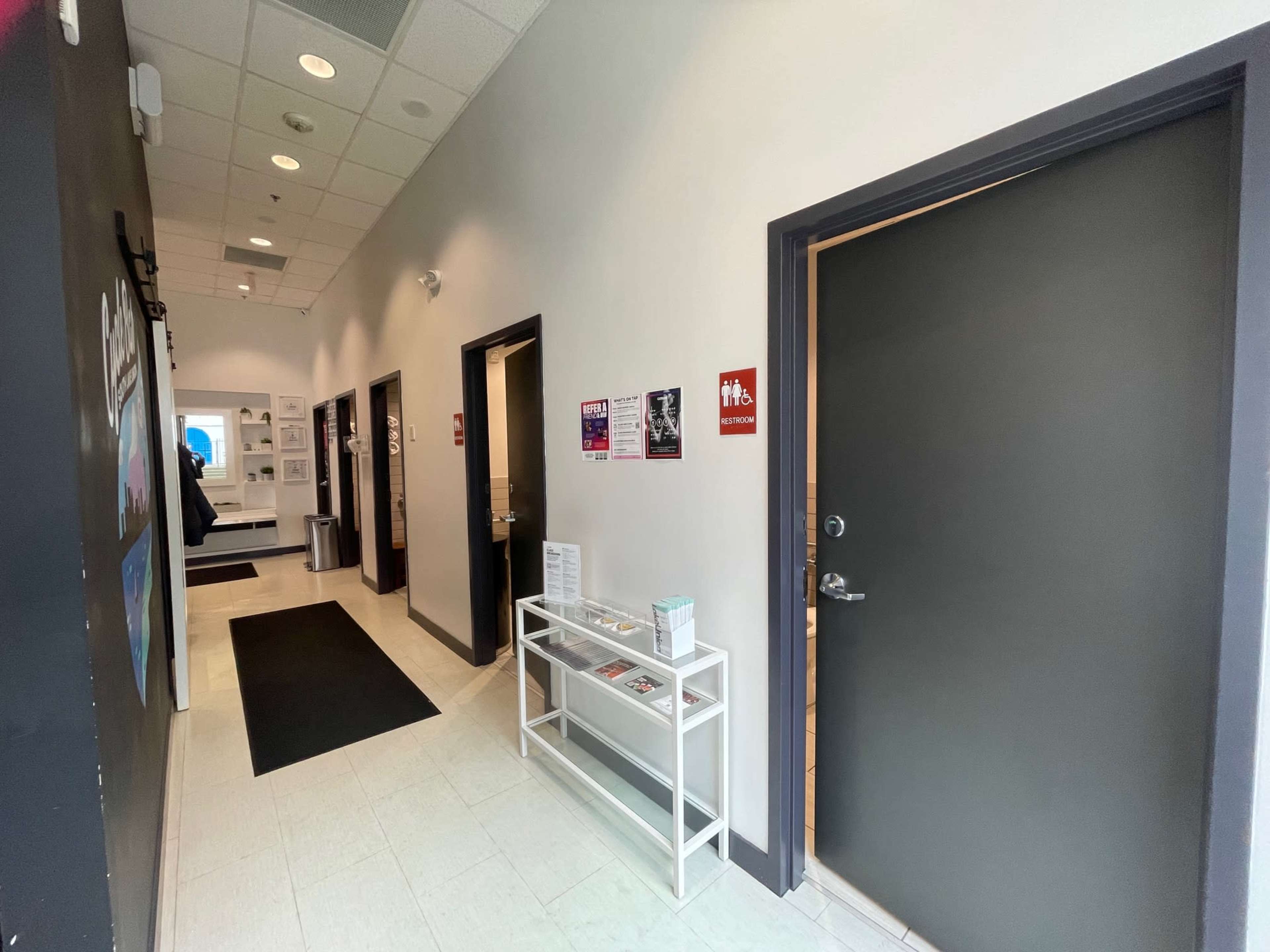 A hallway features doors on the right, with a restroom sign and a white table displaying brochures against a gray wall.